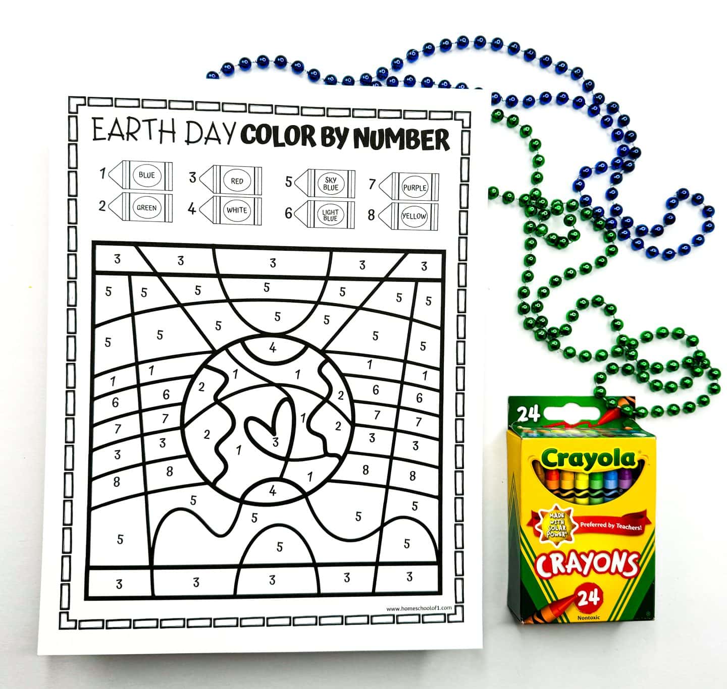 Uncolored Earth Day color by number sheet featuring a heart-shaped Earth in the center. The coloring key lists numbers 1 through 8 paired with specific colors: 1 (blue), 2 (green), 3 (red), 4 (white), 5 (sky blue), 6 (light blue), 7 (purple), and 8 (yellow). A box of 24 Crayola crayons sits to the right, with green and blue beaded garlands above the sheet.