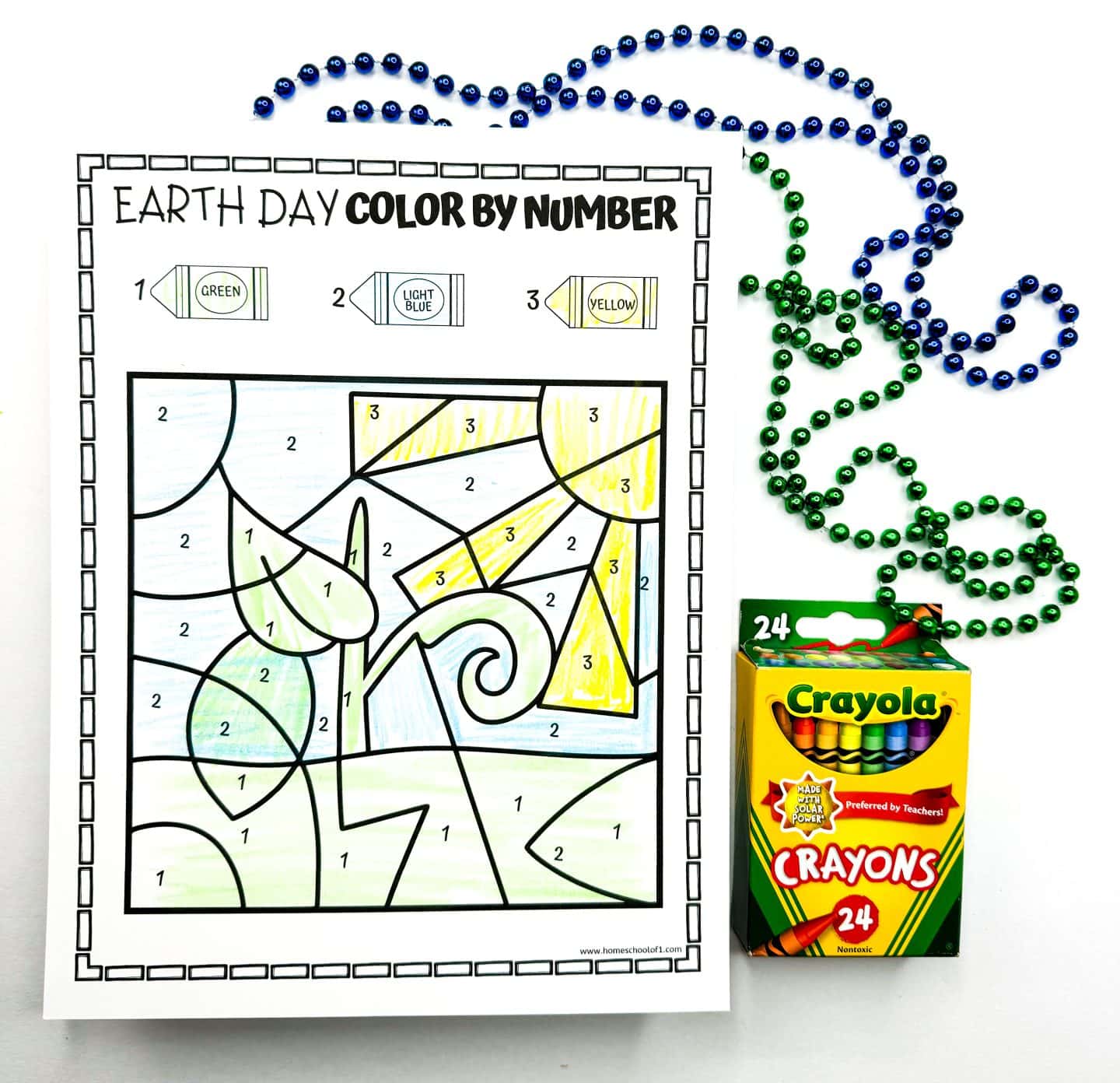Earth Day activity page showing a sprouting plant under a shining sun, filled in using a number-to-color legend. Green, light blue, and yellow crayons are used to complete the vibrant springtime scene. A box of Crayola crayons and colorful bead necklaces sit beside the completed educational coloring sheet.