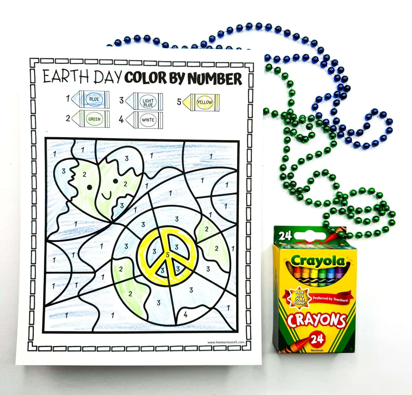 Earth Day color by number worksheet showing a globe with a peace symbol in the center. The coloring key assigns numbers to colors: 1 (blue), 2 (green), 3 (light blue), 4 (white), and 5 (yellow). The earth is shaded in green and blue, with the peace sign in yellow. The background is light blue, with green and blue bead strands and a 24-pack of Crayola crayons beside the sheet.