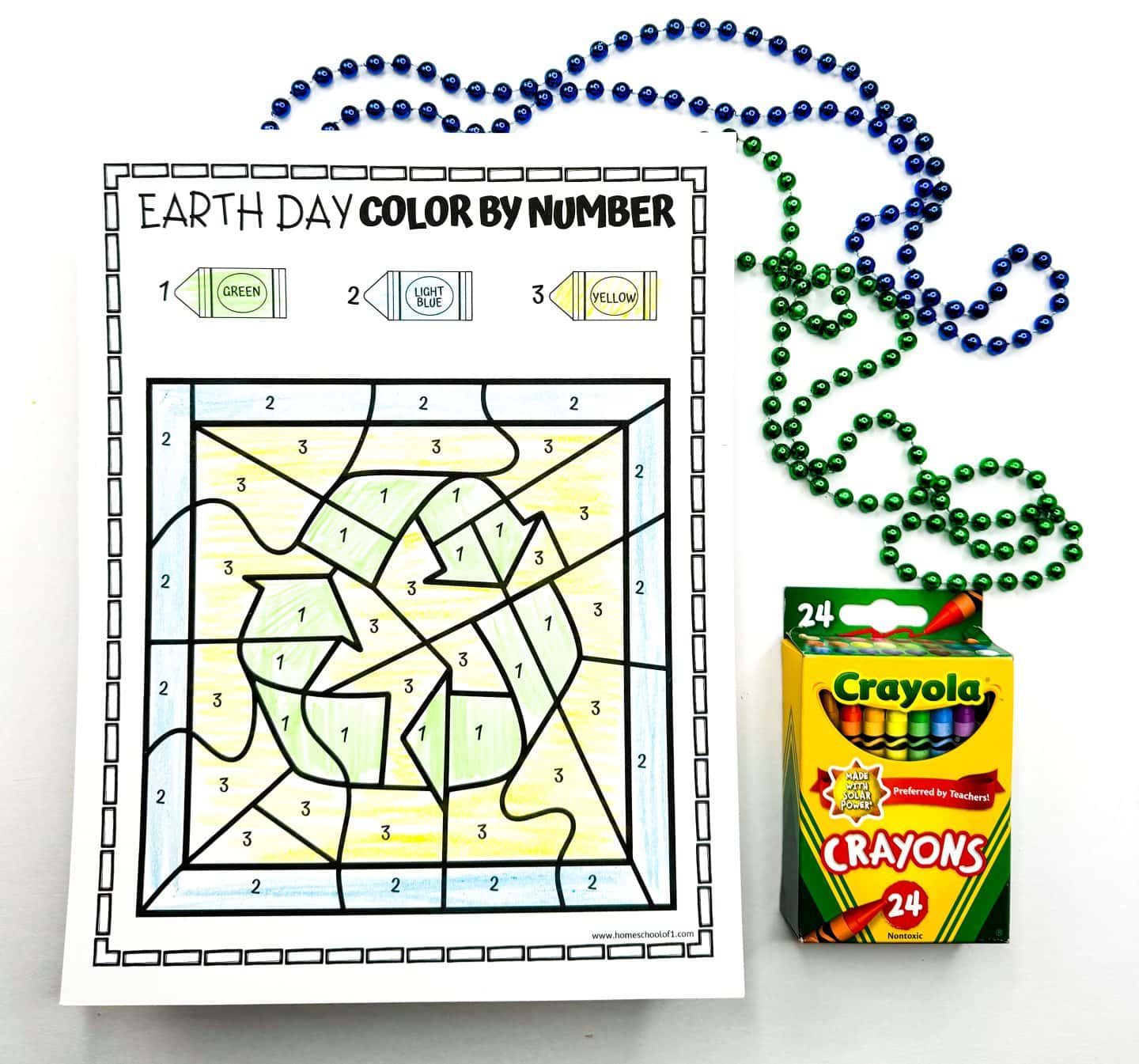 Earth Day color-by-number worksheet featuring a large recycling symbol at the center, filled in with green, set against a background shaded with light blue and yellow. The number key at the top shows 1 for green, 2 for light blue, and 3 for yellow. The worksheet is placed beside green and blue bead strands and a box of 24 Crayola crayons.