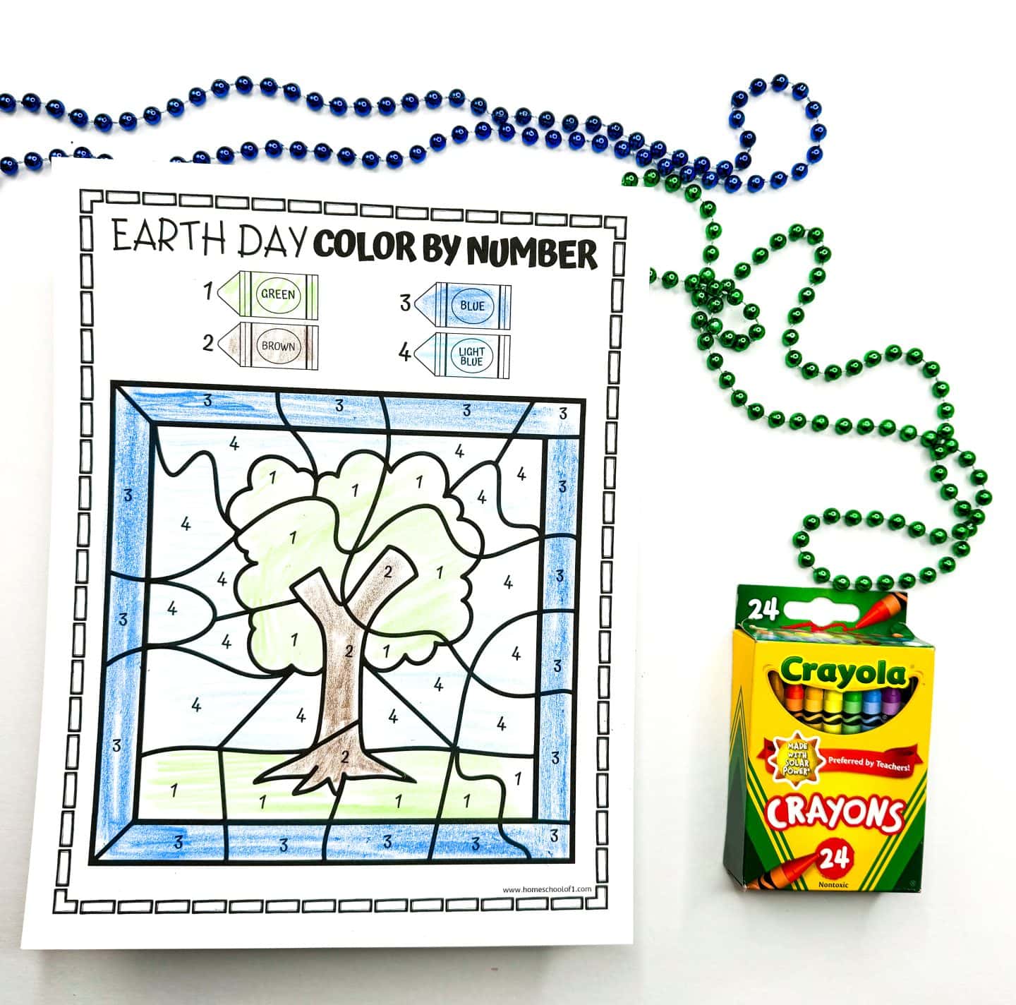 An Earth Day color-by-number worksheet featuring a large tree with green leaves and a brown trunk, set against a background of grass and sky. The coloring guide assigns numbers to colors: green (1), brown (2), blue (3), and light blue (4). The worksheet is surrounded by green and blue beaded necklaces, with a box of 24 Crayola crayons placed on the right side.