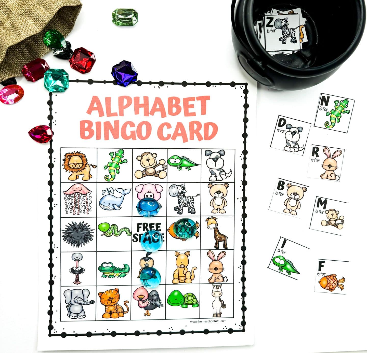  An Alphabet Bingo Board is partially marked with large, colorful gem tokens placed on illustrated animal squares like a hippo, fish, and owl. The grid features playful animals for each letter, such as a lion, jellyfish, turtle, and rabbit. To the right, letter cards like N is for Newt and F is for Fish are spread out near a black bowl filled with more alphabet cards.