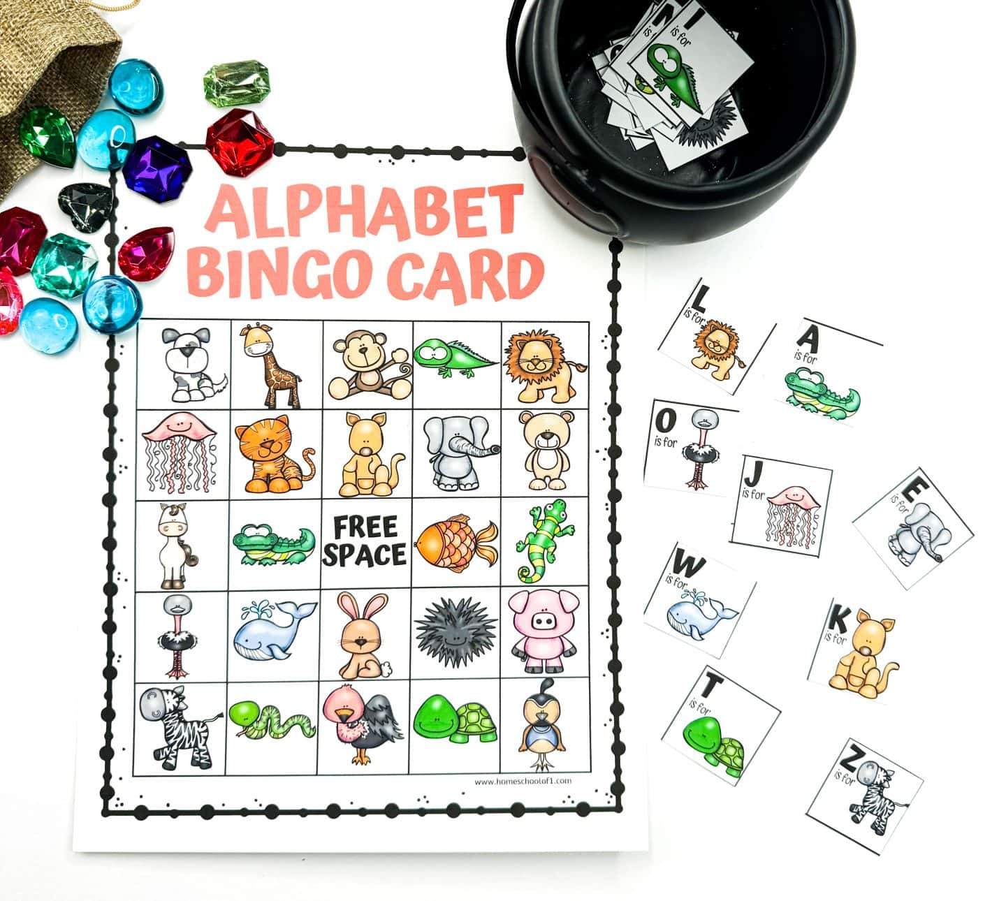 An Alphabet Bingo Card is surrounded by colorful plastic gems and animal-themed letter cards. The bingo grid is filled with cartoon animals like a lion, jellyfish, turtle, and pig, each representing a letter of the alphabet. To the side, loose letter cards including L is for Lion and J is for Jellyfish are laid out near a black bowl containing more cards.