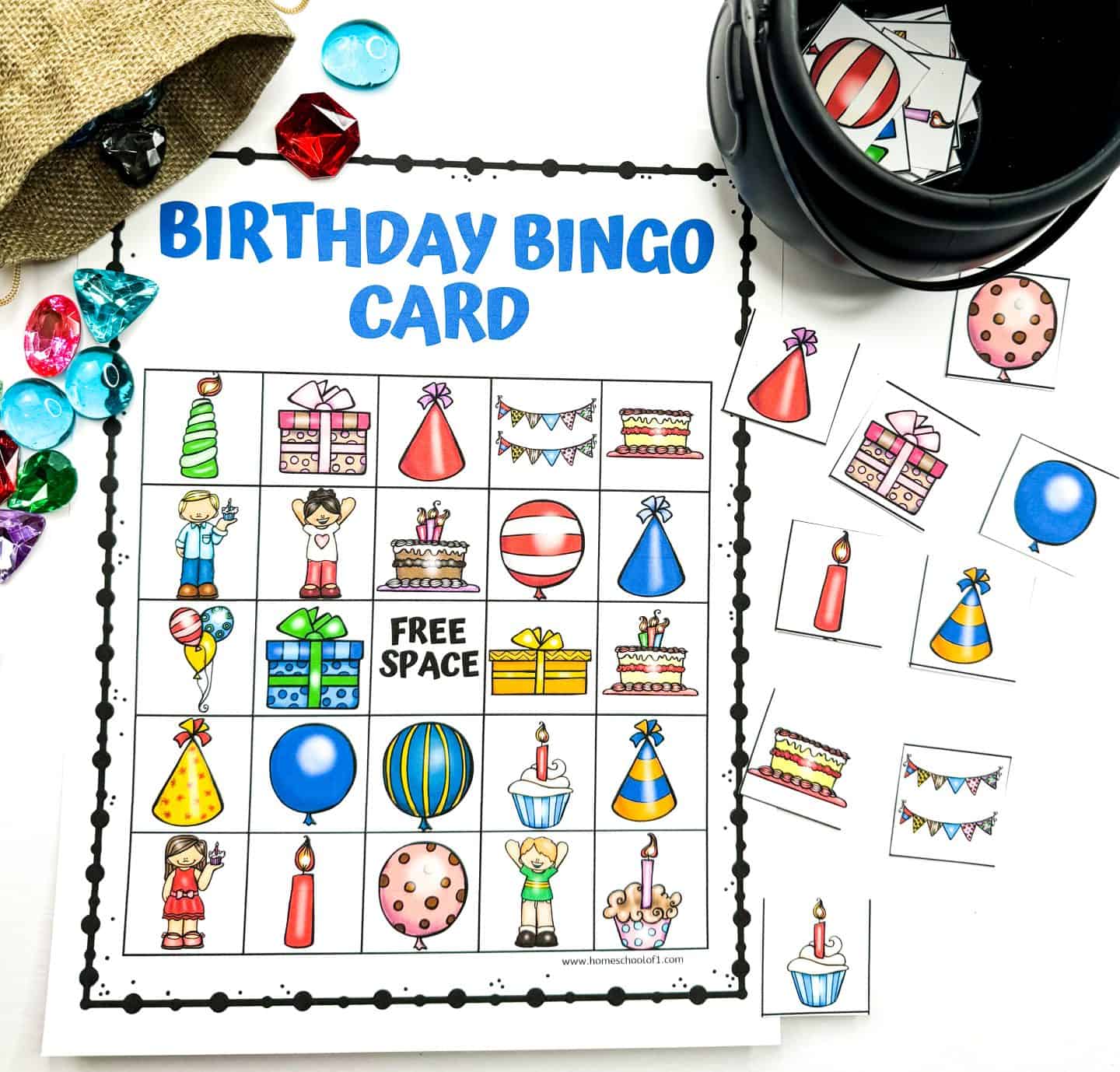 Bright and playful "Birthday Bingo Card" with a 5x5 grid filled with cartoon images like cakes, candles, balloons, presents, cupcakes, party hats, and children celebrating, plus a “FREE SPACE” square. The setup includes colorful gem markers, cut-out bingo pieces, and a black container holding more image tiles, all arranged on a white background.