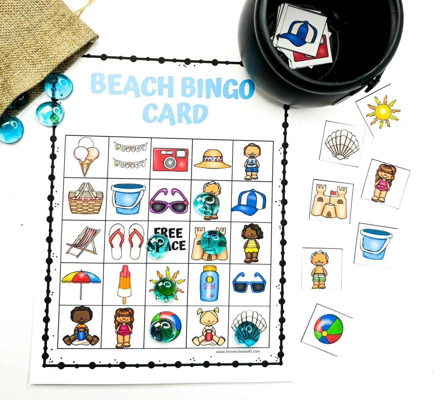 Beach Bingo printable game card filled with colorful beach-themed icons like a sandcastle, sunhat, flip-flops, sunglasses, and ice cream. Several spots are marked with translucent blue glass markers. Surrounding the board are cut-out image tiles, a small burlap bag, and a black container holding more bingo pieces. The title “BEACH BINGO CARD” appears in light blue at the top.

