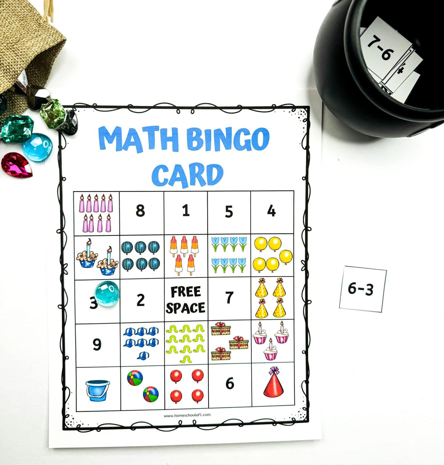 Printable math bingo card for kids featuring colorful illustrations like balloons, cupcakes, party hats, and beach balls in a 5x5 grid. A translucent gem marks one square, and a “6–3” math flashcard sits beside the board. A black cup holds more math problem cards, with additional plastic gems scattered around.