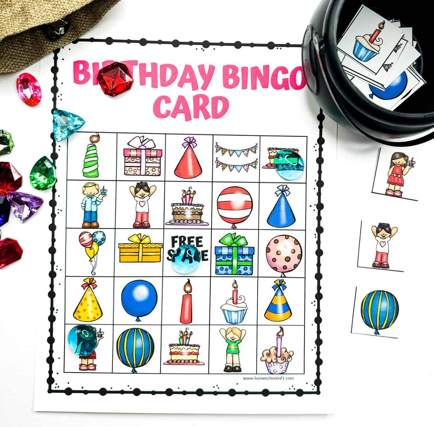 Birthday Bingo card with a pink title and a 5x5 grid filled with colorful icons like balloons, candles, party hats, cupcakes, presents, and children. Some squares are marked with translucent gem tokens, while extra bingo tiles and shiny plastic jewels are scattered around the card and a black container.