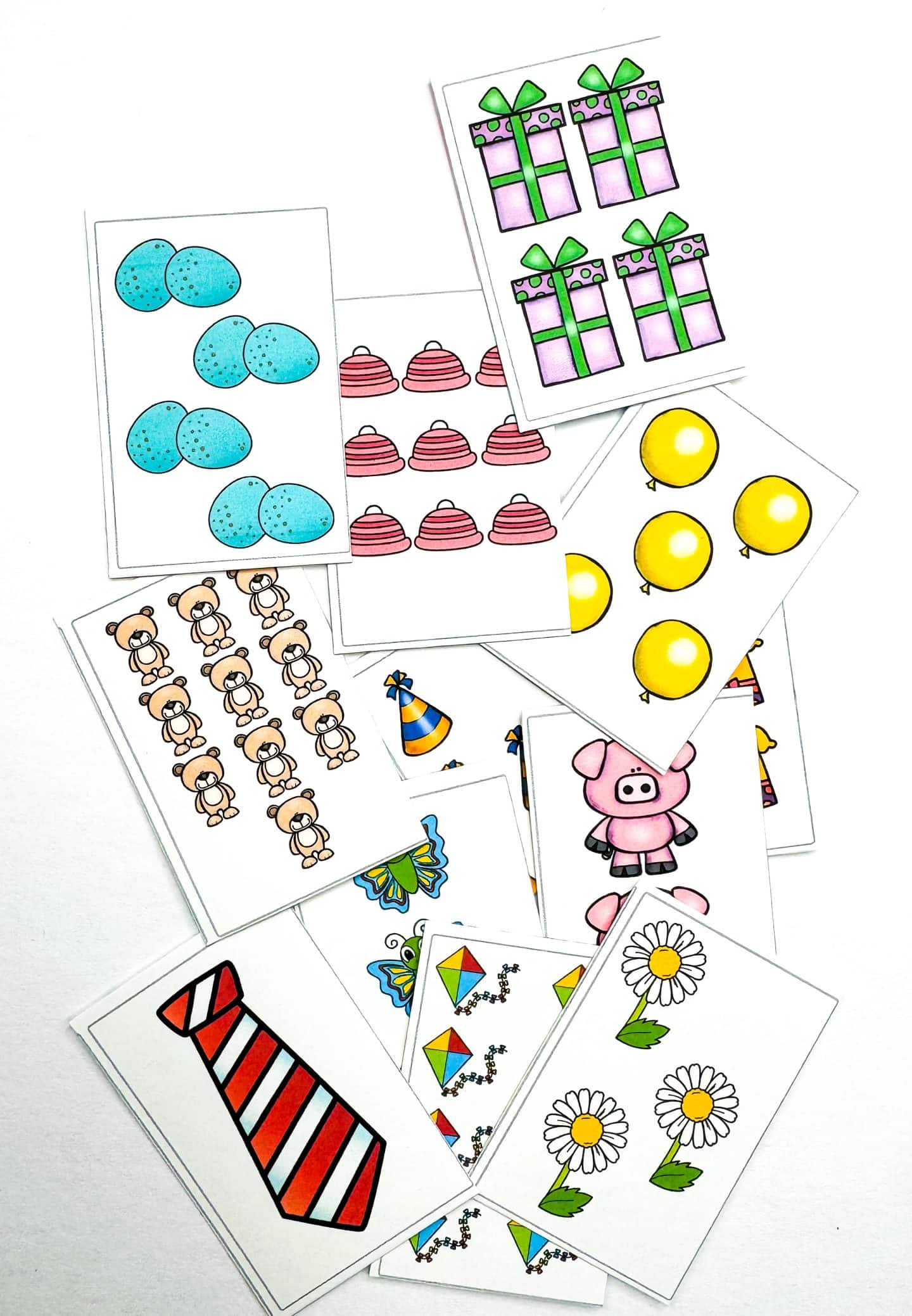 A collection of brightly colored printable memory game cards featuring illustrated objects for counting, such as blue eggs, yellow balloons, pink cupcakes, teddy bears, gifts, party hats, butterflies, kites, daisies, and a striped necktie. These cards are designed to help kids develop number recognition and matching skills through playful visuals.
