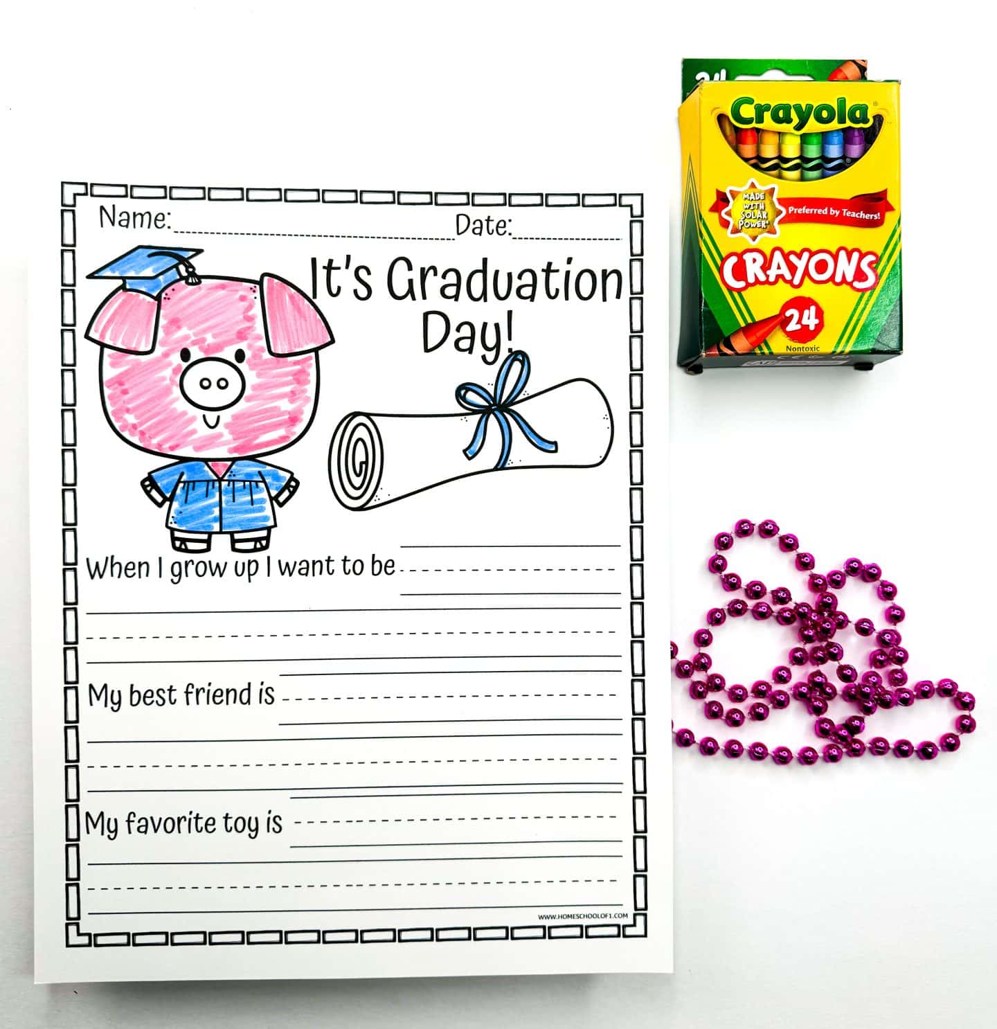 A "It's Graduation Day!" writing prompt worksheet featuring a pink cartoon pig wearing a blue graduation cap and gown, next to a rolled diploma tied with a blue ribbon. The worksheet includes prompts for "When I grow up I want to be," "My best friend is," and "My favorite toy is." A box of Crayola crayons and a string of shiny purple beads are placed beside the worksheet.