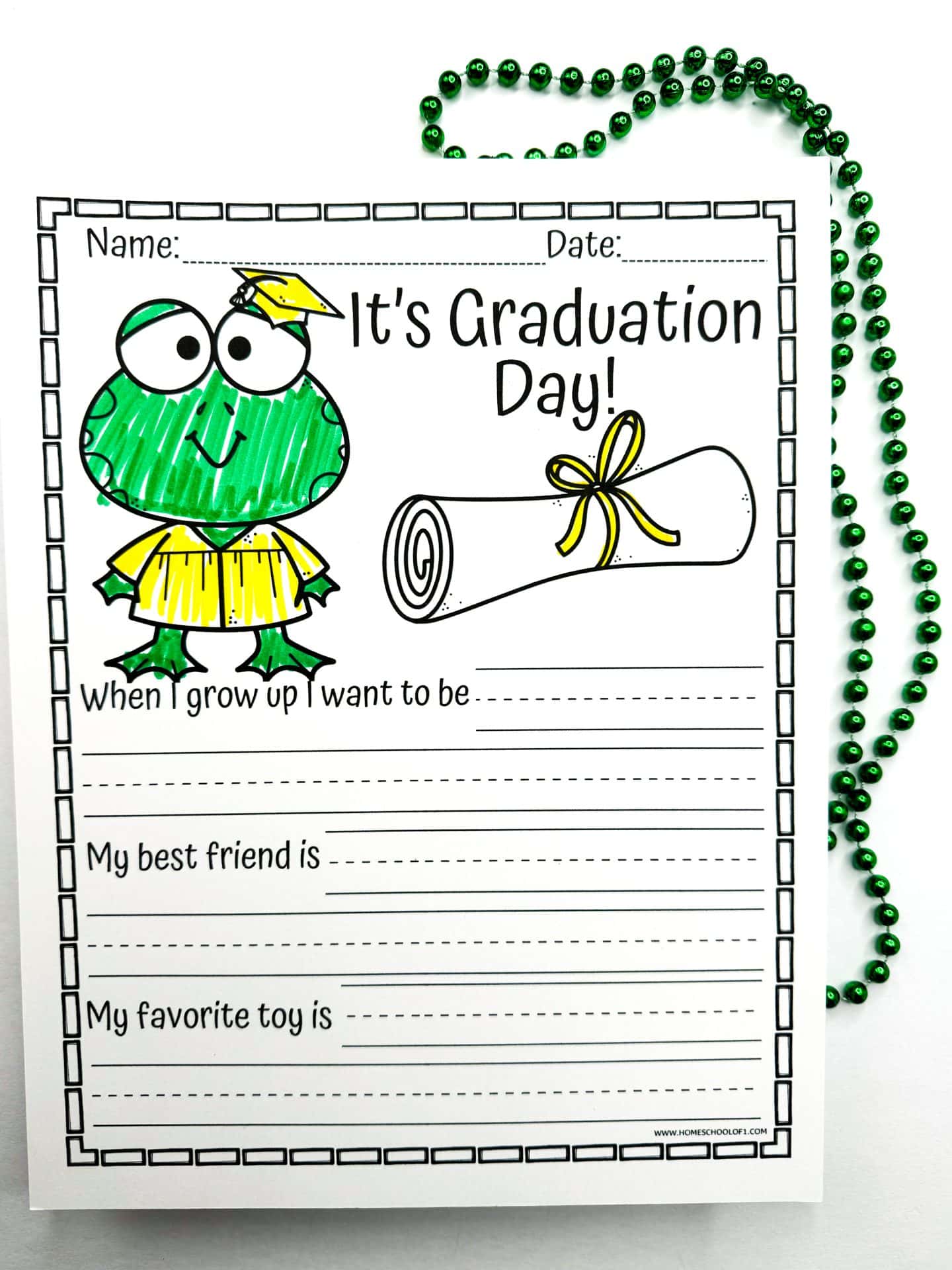 A "It's Graduation Day!" writing prompt worksheet featuring a green cartoon frog wearing a yellow graduation cap and gown, beside a rolled diploma tied with a yellow ribbon. The worksheet has sections for kids to write answers to "When I grow up I want to be," "My best friend is," and "My favorite toy is," and is decorated with a string of shiny green beads.