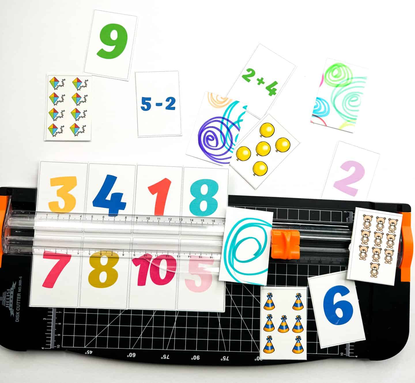 Colorful math memory game cards in the process of being cut with a paper trimmer. Cards feature bold numbers, math problems like “2+4” and “5-2,” and images such as balloons, kites, and party hats for counting practice. A grid paper cutting mat is underneath, and swirly-patterned backs add a fun visual element.