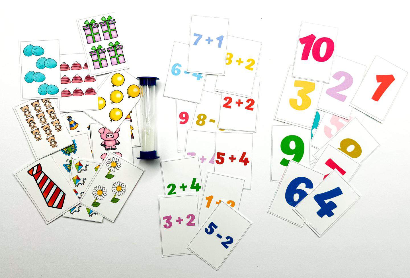 Colorful math memory game cards scattered across a white surface, featuring numbers, simple addition and subtraction problems, and illustrated object sets like balloons, cakes, and flowers. A sand timer is placed in the center, suggesting timed play for extra challenge. Cards display bold colors and visuals to support number recognition and counting skills.