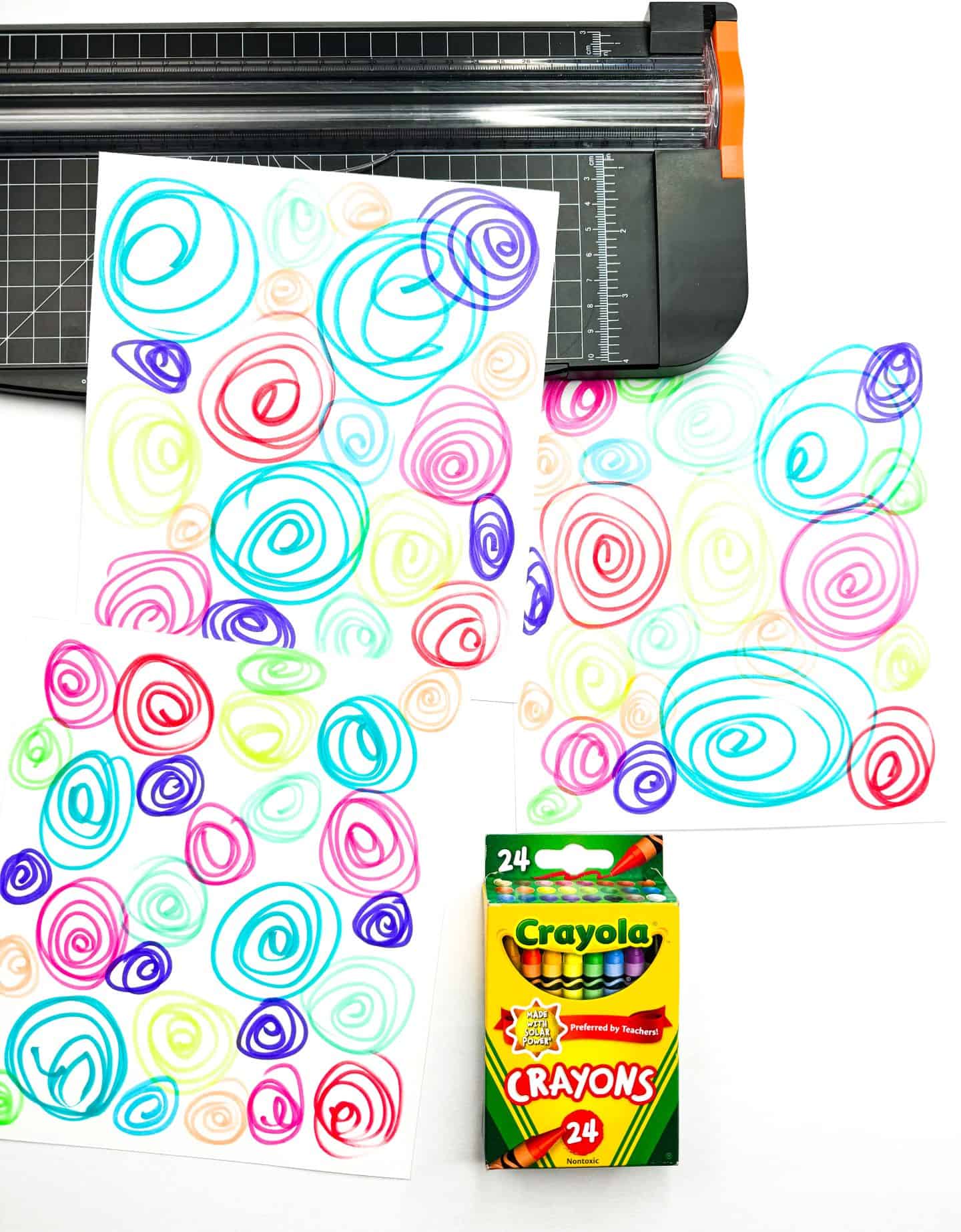 Two brightly colored sheets covered in hand-drawn spiral doodles in a variety of vibrant colors lie next to a black paper trimmer. A box of 24 Crayola crayons is placed in the lower right corner, suggesting the materials were used to create the artwork.