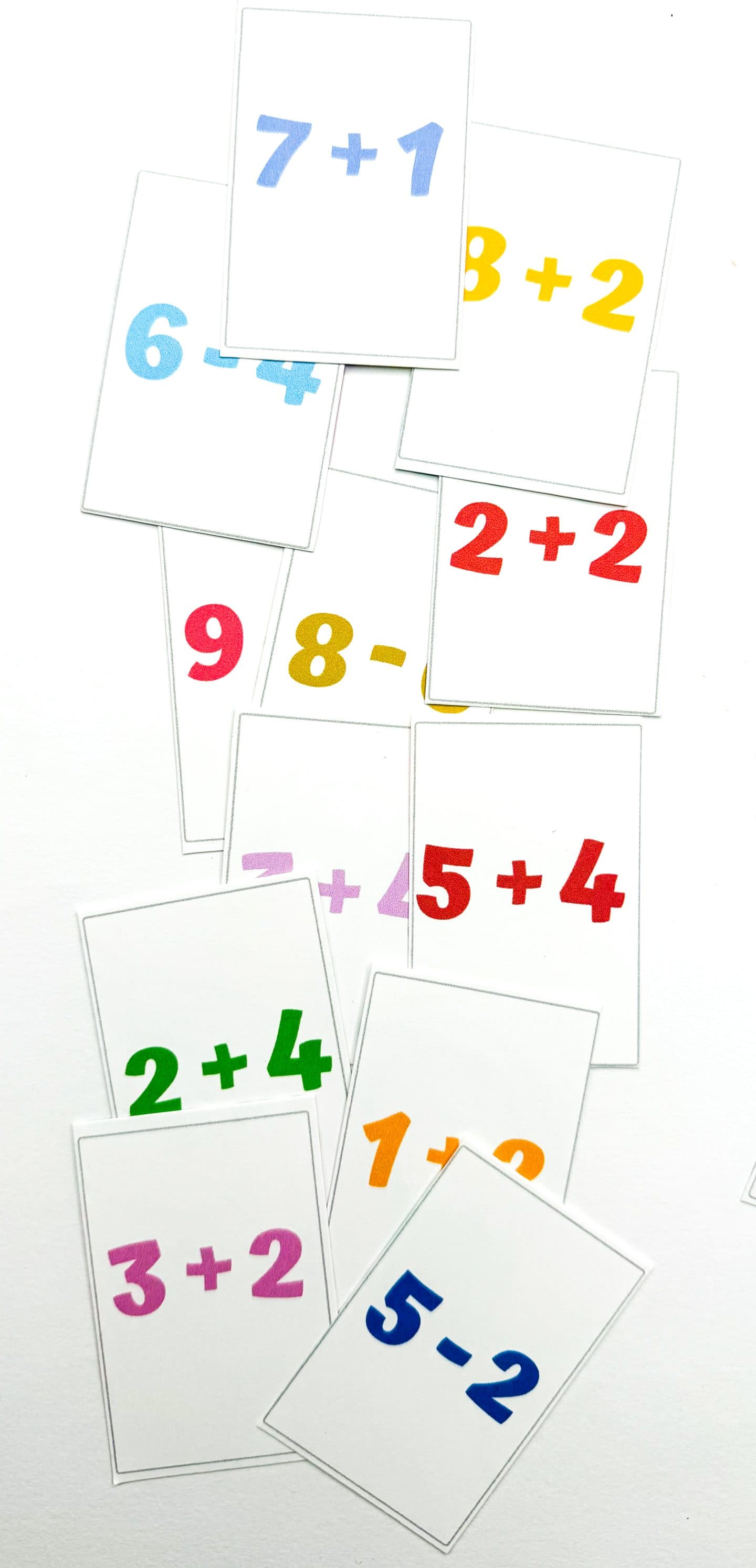 A scattered collection of printable flashcards showing colorful basic math equations like 7+1, 2+2, 5-2, 3+2, and 8-0. Each card features large, bold numbers and symbols in different colors on a white background, designed to help young learners practice addition and subtraction in a fun, hands-on way.