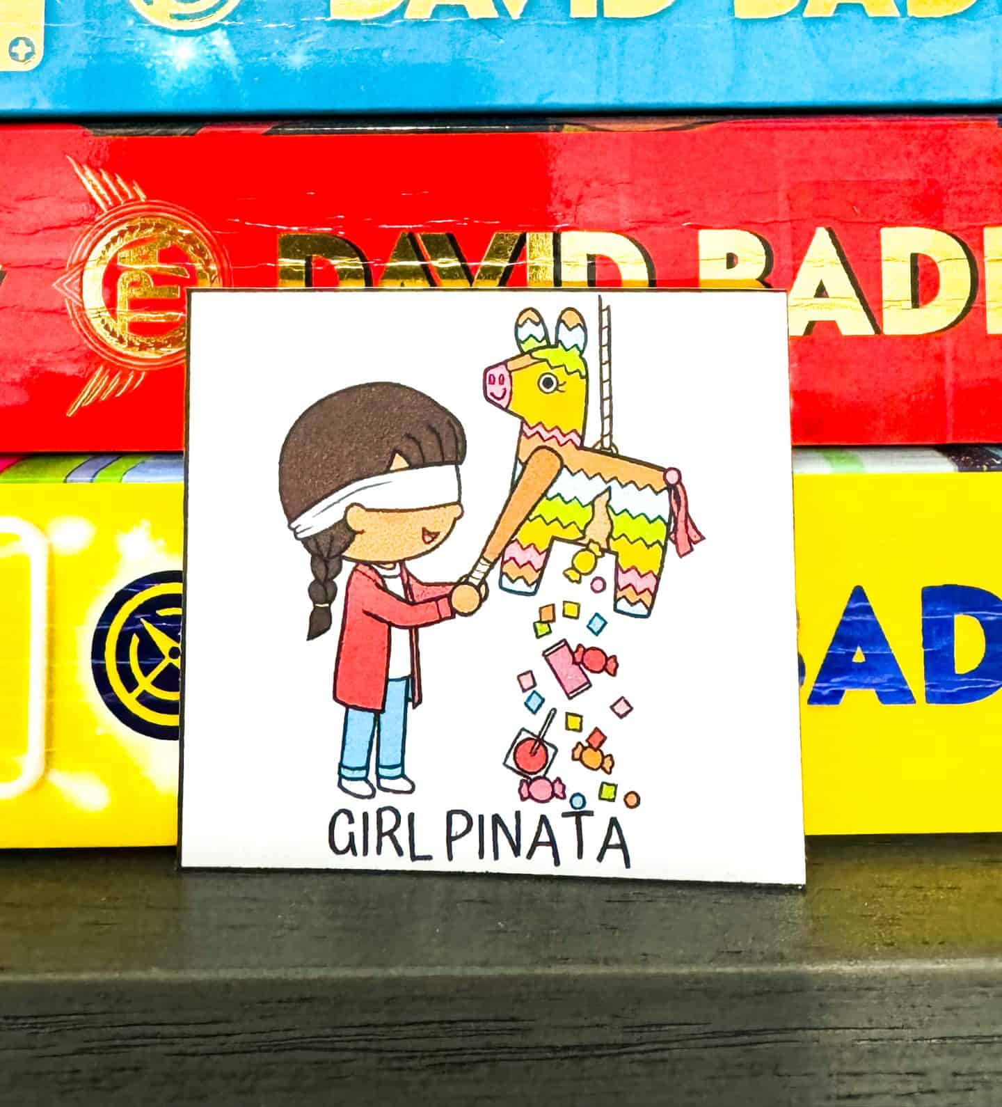 A colorful illustration from a 'Cinco de Mayo Scavenger Hunt' of a blindfolded girl in red hitting a festive multicolored piñata, with candy spilling out, placed on a surface with books in the background.