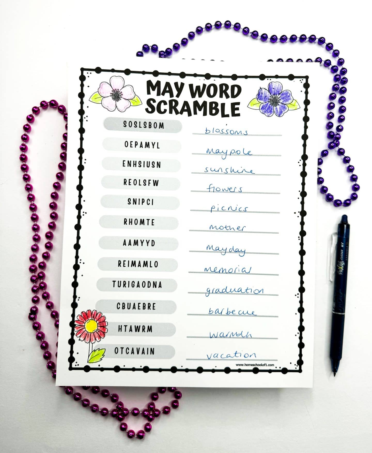 Challenging May Word Scramble sheet with scrambled words like 'blossoms' and 'vacation' alongside their solved counterparts, surrounded by Mardi Gras beads and a blue pen