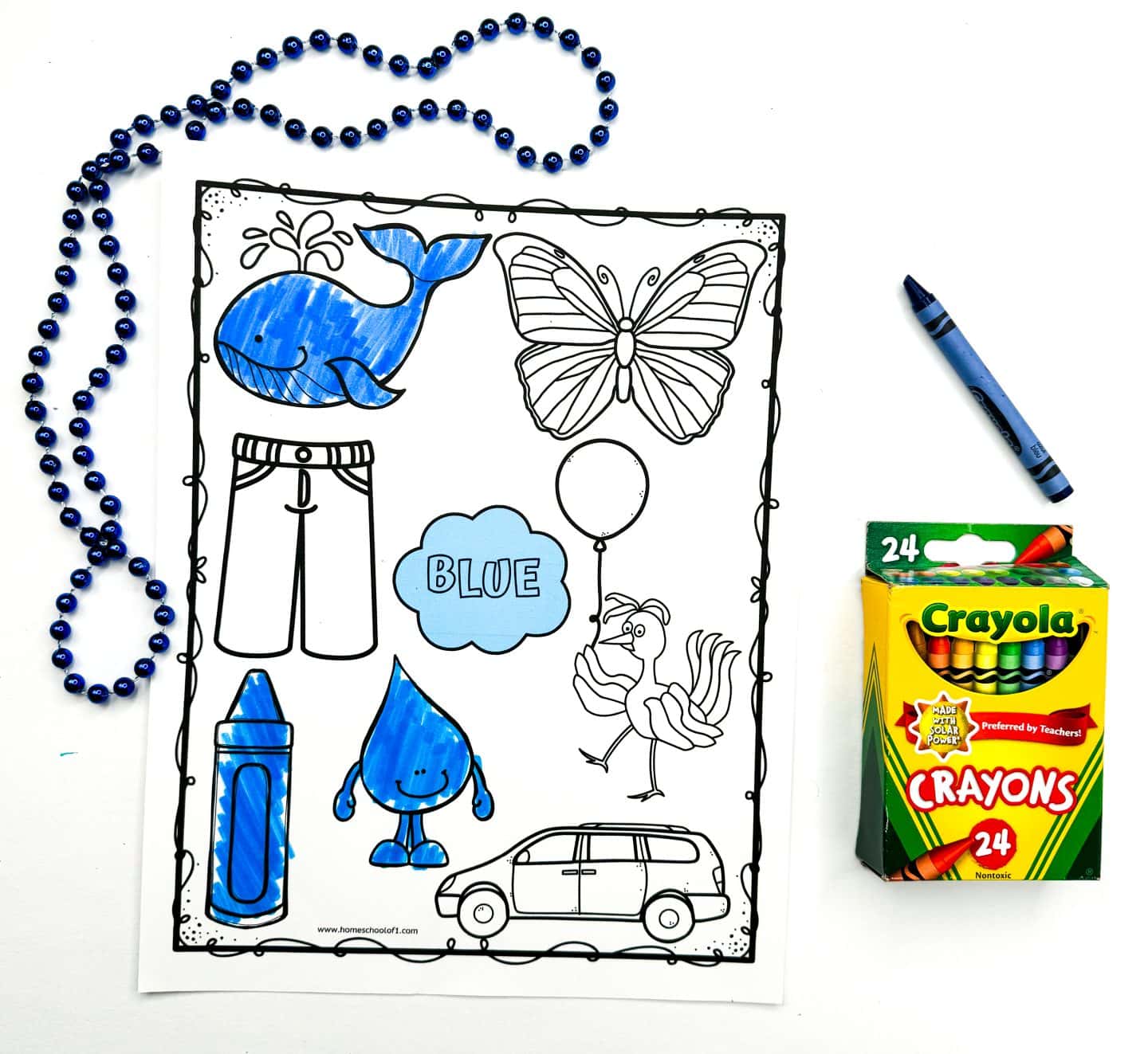 A blue coloring worksheet includes outlined images such as a whale, butterfly, crayon, water droplet, and a cloud labeled blue, with several items partially colored in with a blue crayon. A strand of shiny blue beads lies to the side, and a 24-count Crayola crayon box sits nearby. The worksheet helps children identify and color objects associated with the color blue.