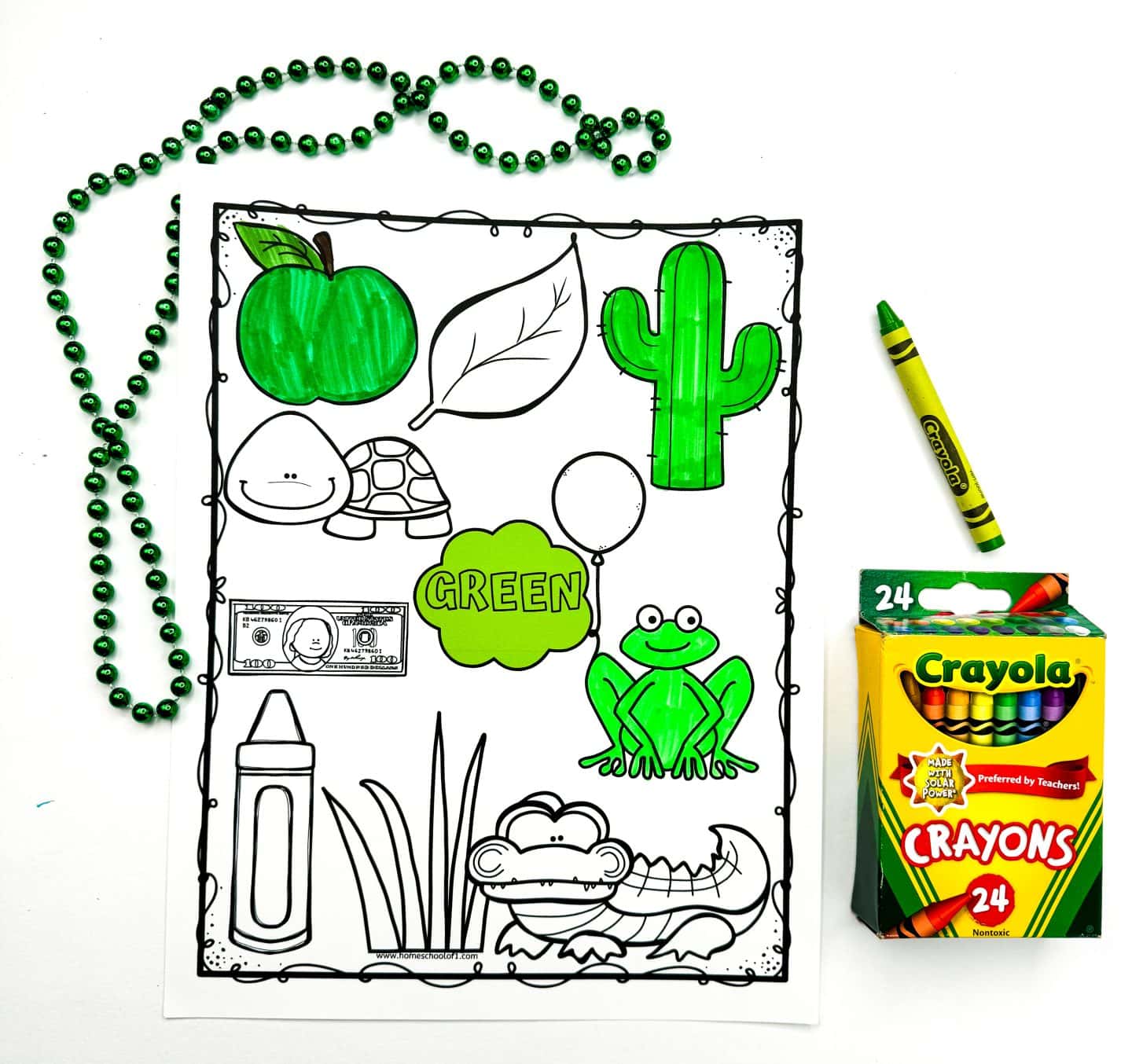 A green-themed printable coloring worksheet features outlined images like an apple, cactus, frog, crayon, balloon, and a cloud labeled green, with several items partially or fully colored in green. A green beaded necklace, green Crayola crayon, and a box of 24 Crayola crayons are arranged around the sheet. The worksheet is designed for early learners to practice color recognition.