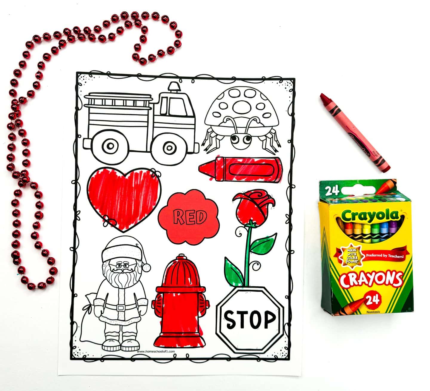 A red coloring worksheet displays outlined images like a fire truck, crayon, heart, rose, fire hydrant, and a cloud labeled red, with several items partially colored in using a red crayon. A red beaded necklace is draped around the page, and a 24-count box of Crayola crayons sits nearby. The worksheet is designed to help kids learn color recognition through engaging illustrations.