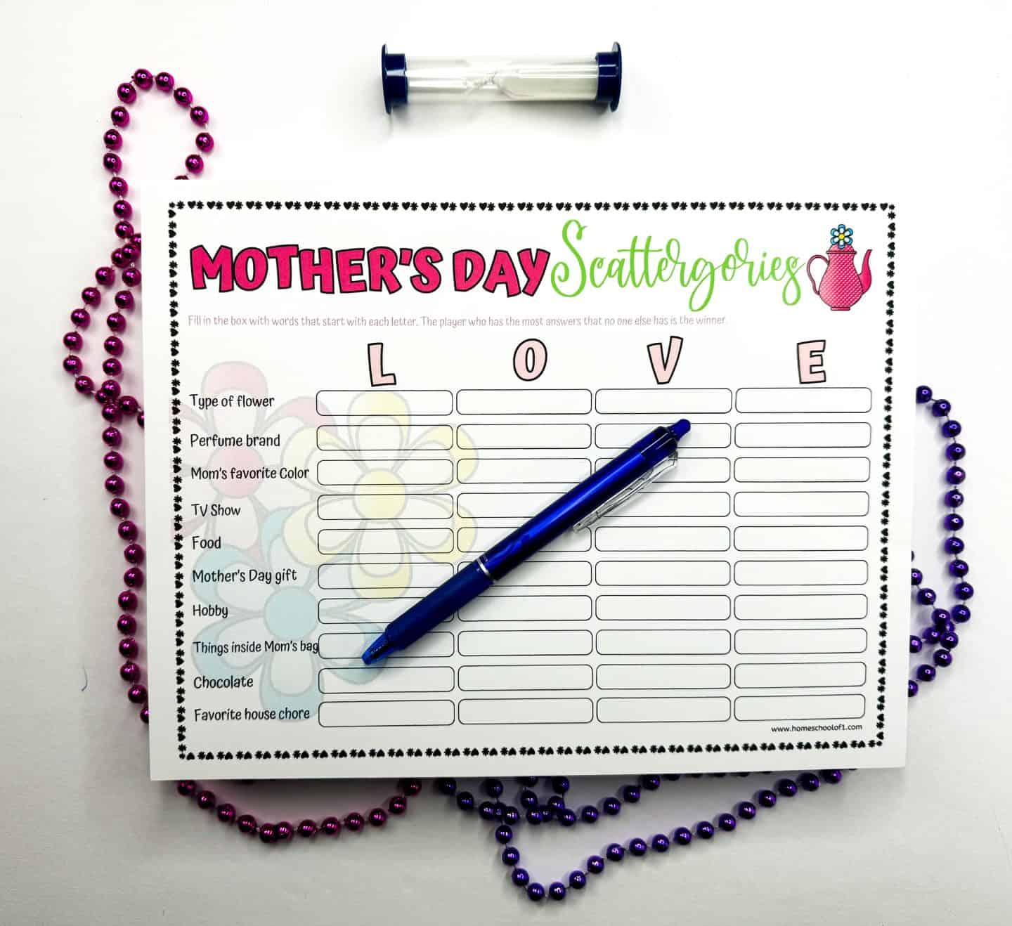 Flat lay of a “LOVE” themed Mother's Day Scattergories game board with a blue pen placed diagonally across the sheet. The categories include type of flower, perfume brand, mom’s favorite color, TV show, food, and more. A sand timer and strands of pink and purple beads frame the scene on a white background.

