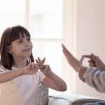 This image captures a tender moment between a young girl with shoulder-length hair and an adult, likely engaging in a sign language lesson. The child, dressed in a simple white shirt, focuses intently on forming hand signs, mirroring the gestures of the adult whose hands are visible in the frame. They are seated in a warmly lit room with soft natural light filtering through a window, emphasizing the serene and focused atmosphere of their learning experience.