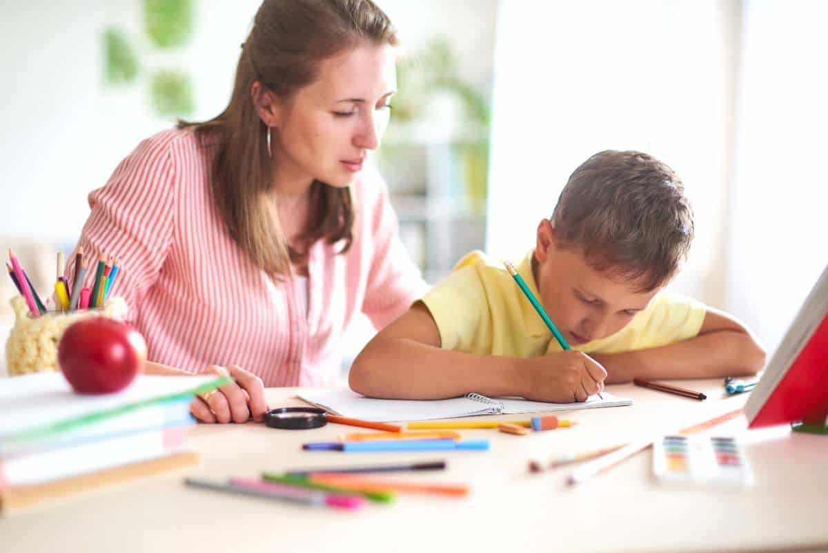 Mother homeschooling her child, sitting at a table with various school supplies, as the child concentrates on writing in a notebook, showcasing a dedicated and personalized learning environment.
