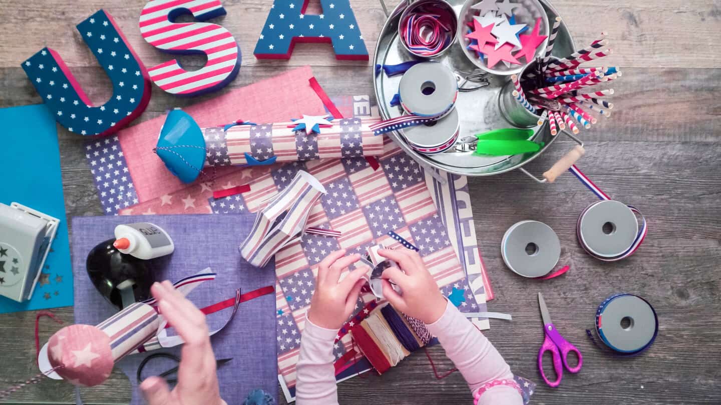 Child’s hands assembling patriotic decorations with red, white, and blue stars, ribbons, paper, and glue for a holiday craft project.