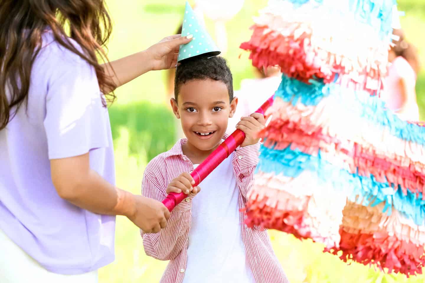 Smiling boy holding a bat next to a colorful piñata while an adult places a polka-dot party hat on his head.