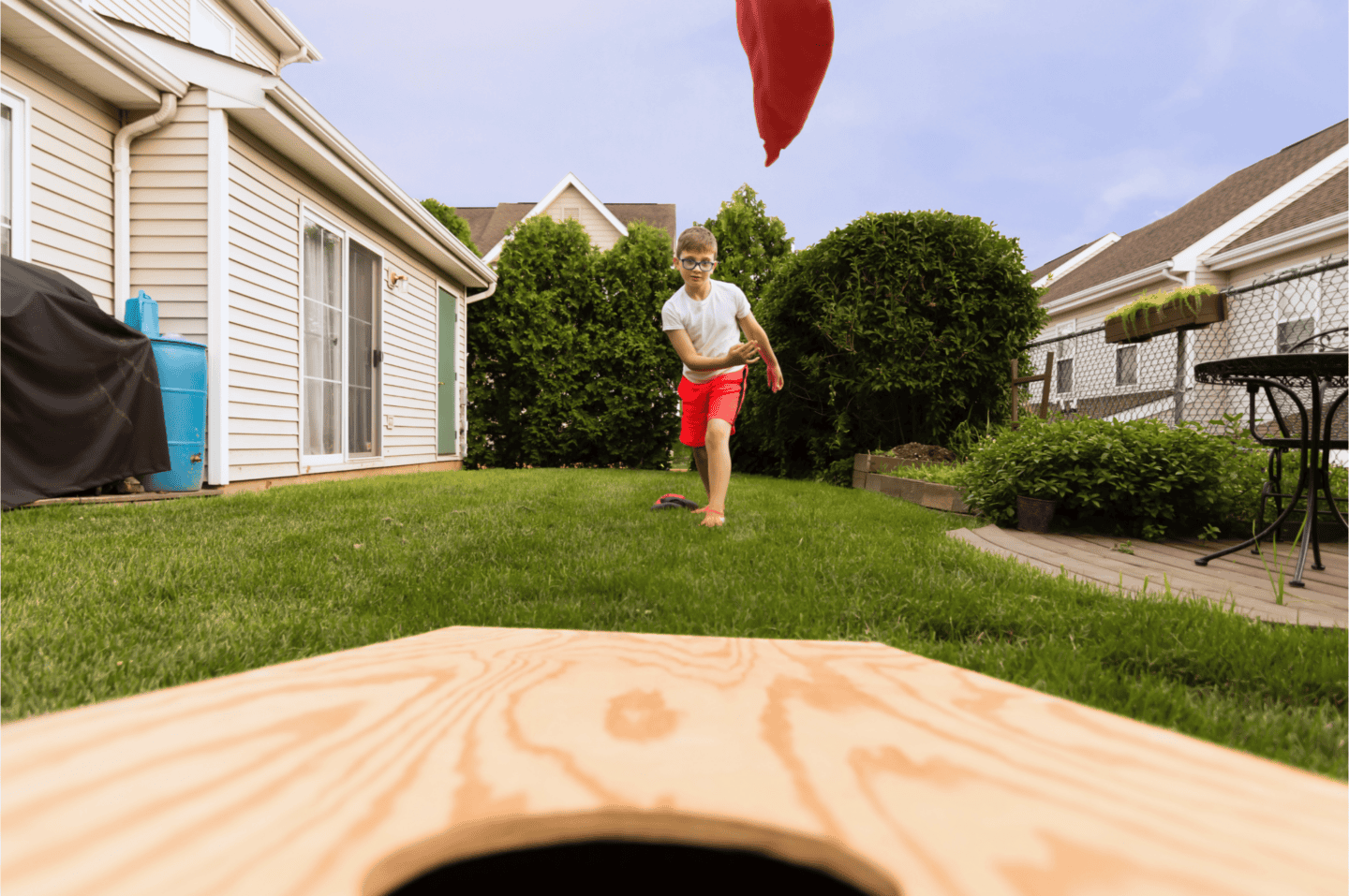 Boy in red shorts and glasses tossing a red bean bag toward a wooden cornhole board in a backyard setting.