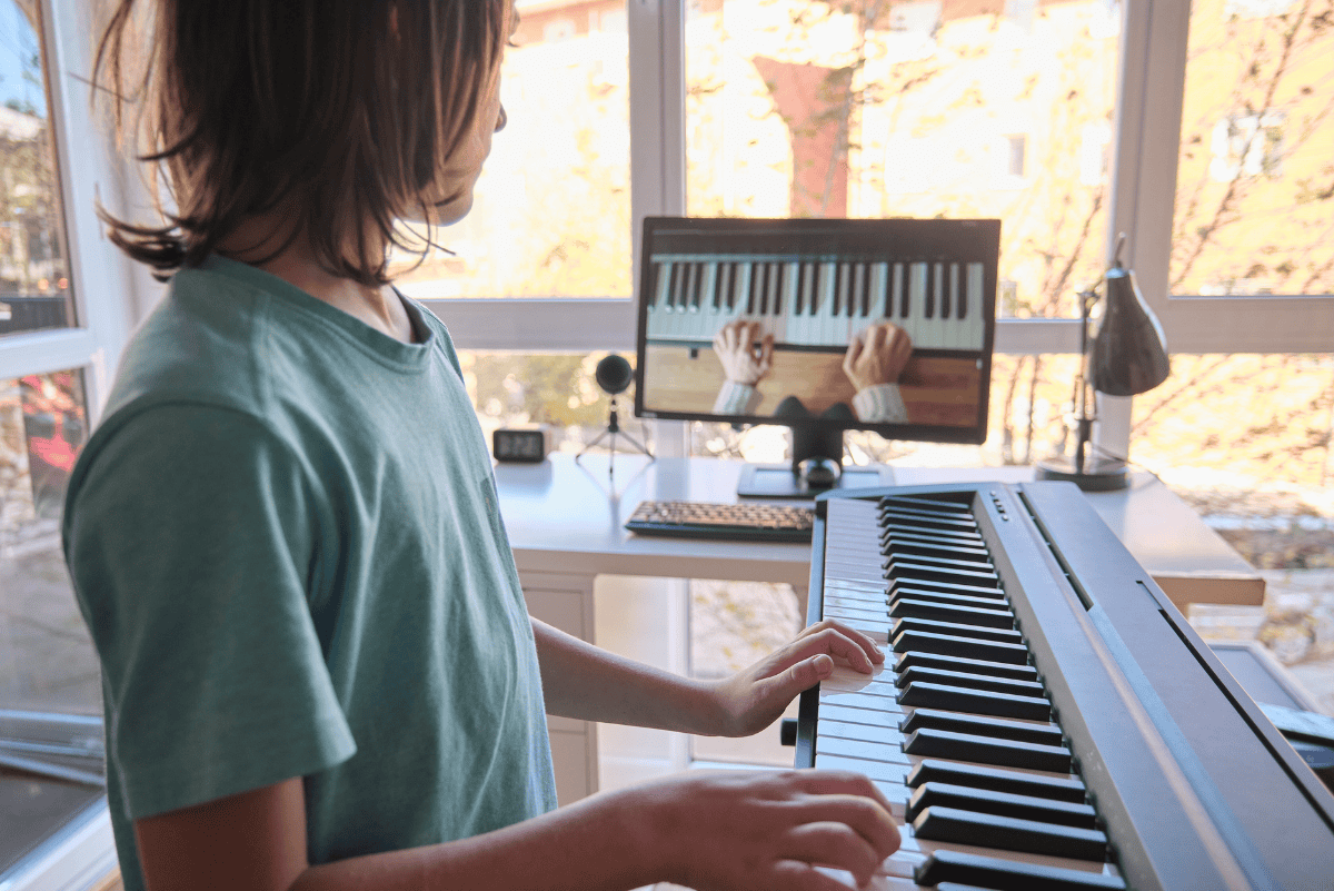 Child in a teal shirt practices on a digital piano while following an online piano lesson displayed on a computer monitor, which shows hands playing keys from an overhead view. Set in a bright, sunlit room, this image highlights a modern and effective setup for the best online piano lessons for kids.