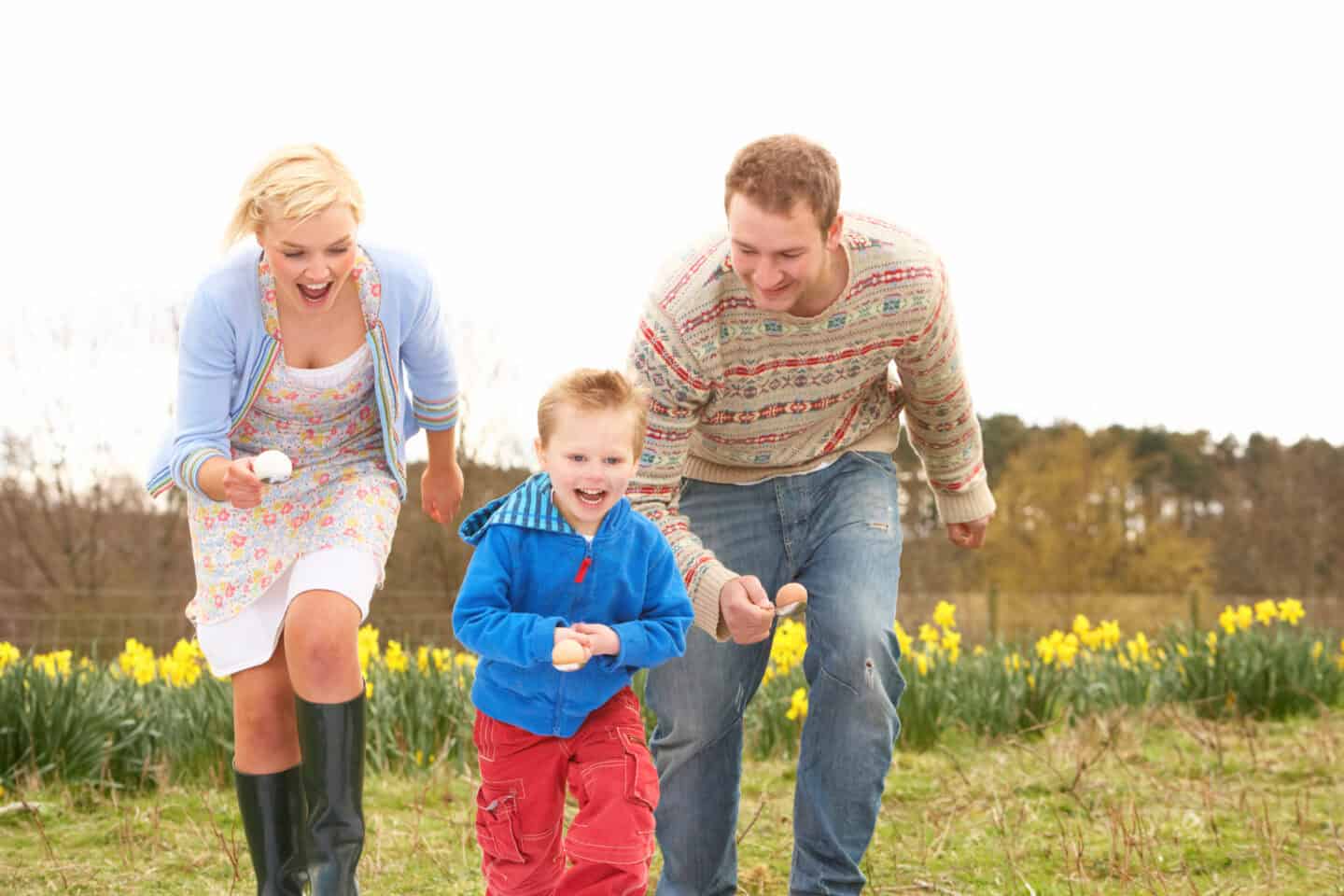 Laughing family of three racing outdoors while balancing eggs on spoons during a springtime event.