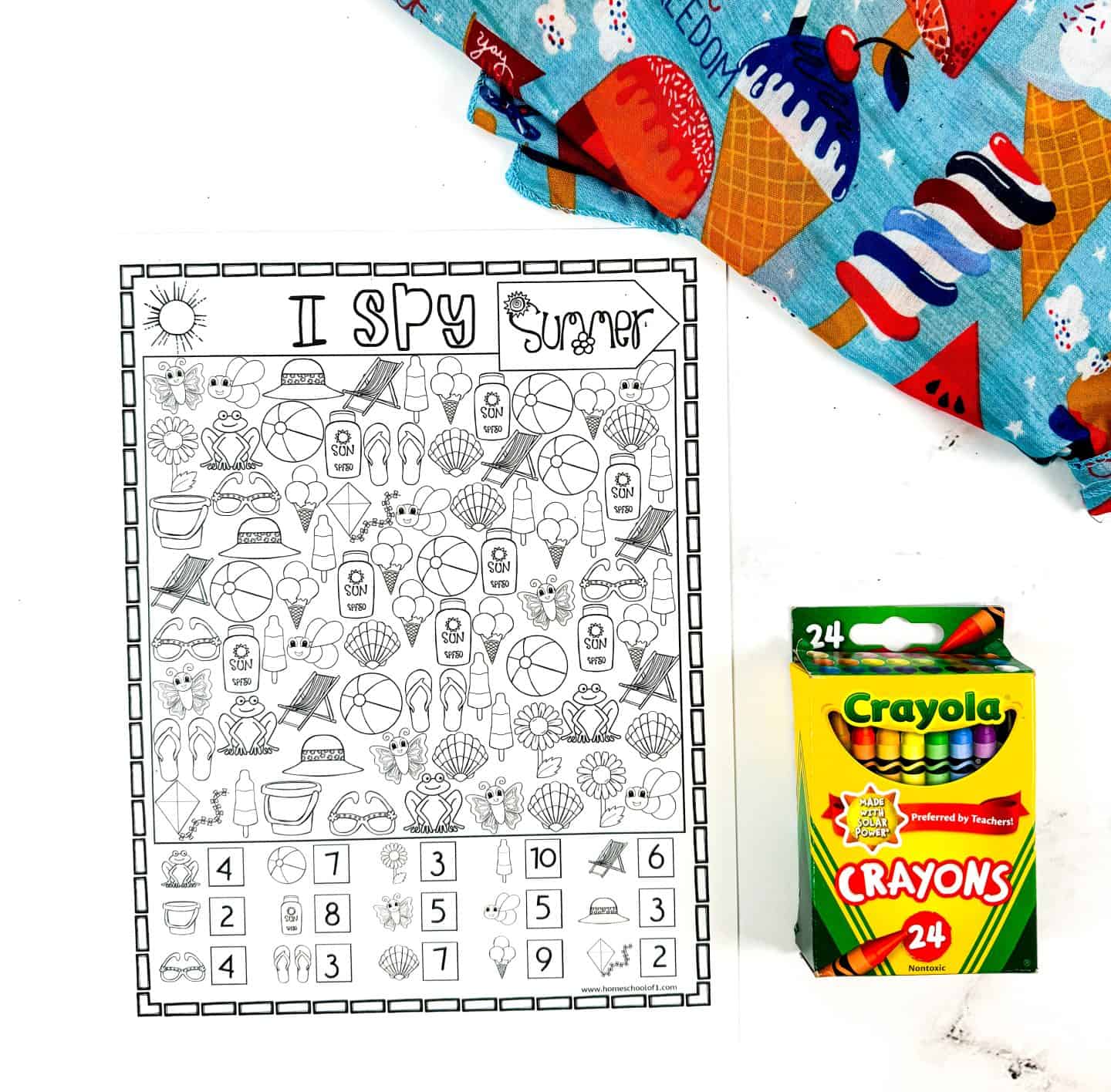 Black-and-white "I Spy Summer" printable featuring a grid of summer-themed icons like beach balls, sunscreen, sunglasses, flip flops, ice cream, and seashells, alongside a count-and-find activity with corresponding numbers at the bottom. The sheet is placed next to a 24-pack of Crayola crayons and a colorful fabric printed with ice cream cones and popsicles.