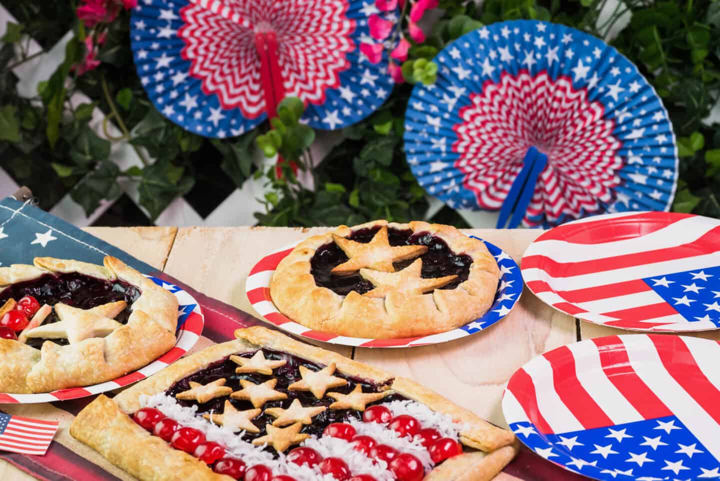 Assorted star-topped berry pies and cherry-flag tart on red, white, and blue star-spangled plates, set with patriotic paper fans in the background.