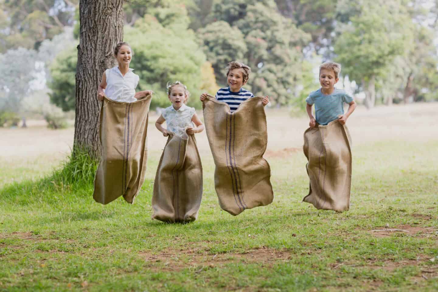 Four children mid-jump in a burlap sack race outdoors on a sunny day.