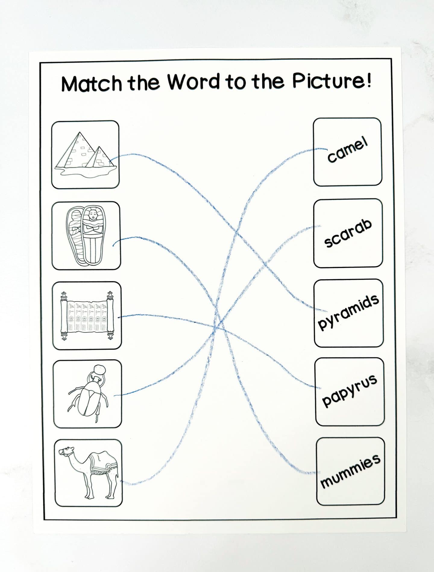 A black-and-white worksheet titled “Match the Word to the Picture!” showing six ancient Egypt-themed illustrations on the left—pyramids, mummies, papyrus scroll, scarab beetle, and a camel—each matched with corresponding words on the right using blue crayon lines: camel, scarab, pyramids, papyrus, and mummies.

