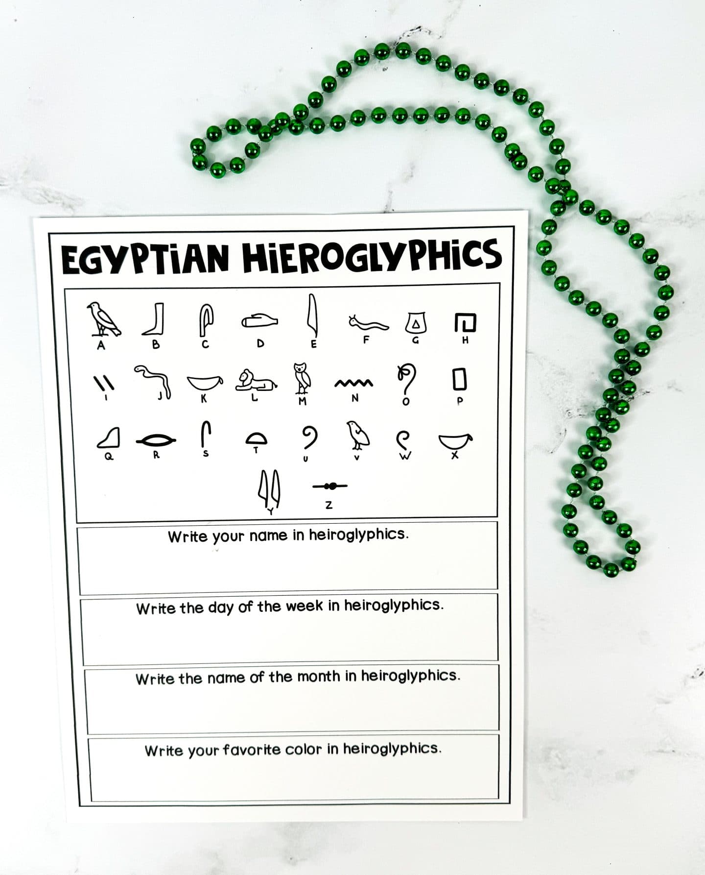 A printable worksheet titled “Egyptian Hieroglyphics” displaying a full A–Z chart of hieroglyphic symbols paired with corresponding English letters. Below the chart are four writing prompts: “Write your name in hieroglyphics,” “Write the day of the week in hieroglyphics,” “Write the name of the month in hieroglyphics,” and “Write your favorite color in hieroglyphics.” A green beaded necklace is placed at the top for decoration.

