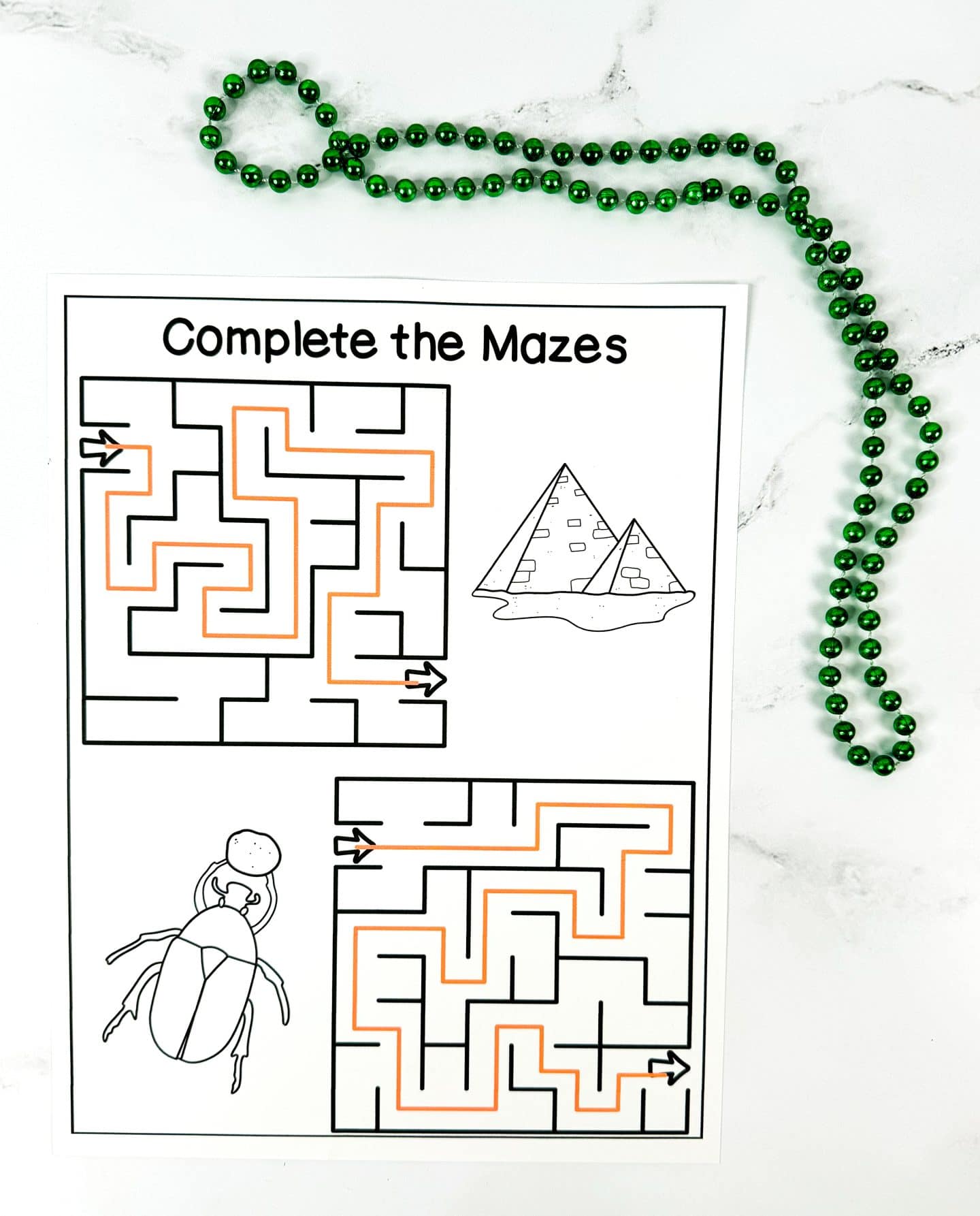 A printable worksheet titled “Complete the Mazes” featuring two simple mazes outlined in black, each with a start and finish arrow. One maze is next to a drawing of pyramids, and the other is next to a large scarab beetle illustration. Both mazes are partially completed with orange crayon lines. A green beaded necklace is placed decoratively at the top.

