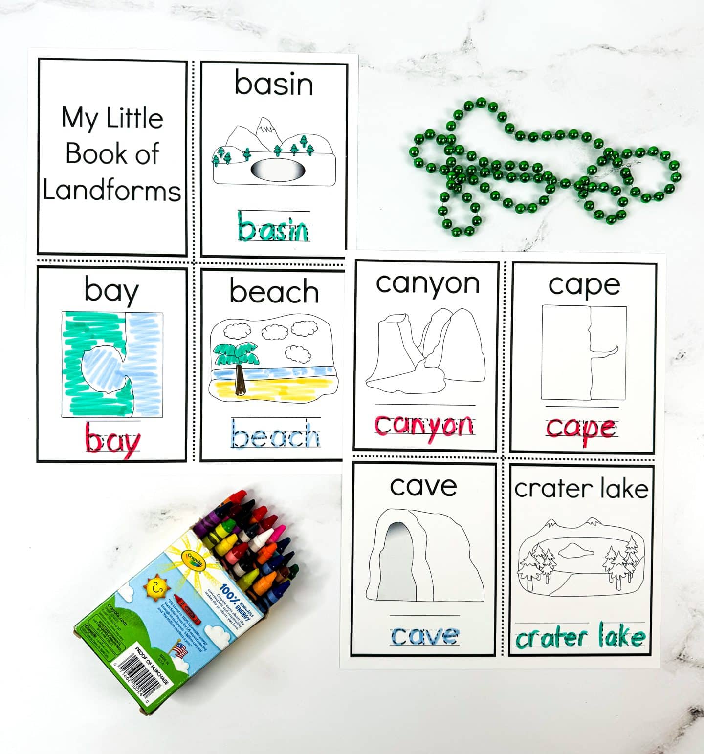 This image displays a set of printable landform flashcards labeled “My Little Book of Landforms.” Each card includes a simple illustration, the landform name in bold, and the name traced in color. The landforms shown are: basin, bay, beach, canyon, cape, cave, and crater lake. A box of crayons and a string of green beads appear beside the cards, suggesting a fun and interactive learning activity for kids.

