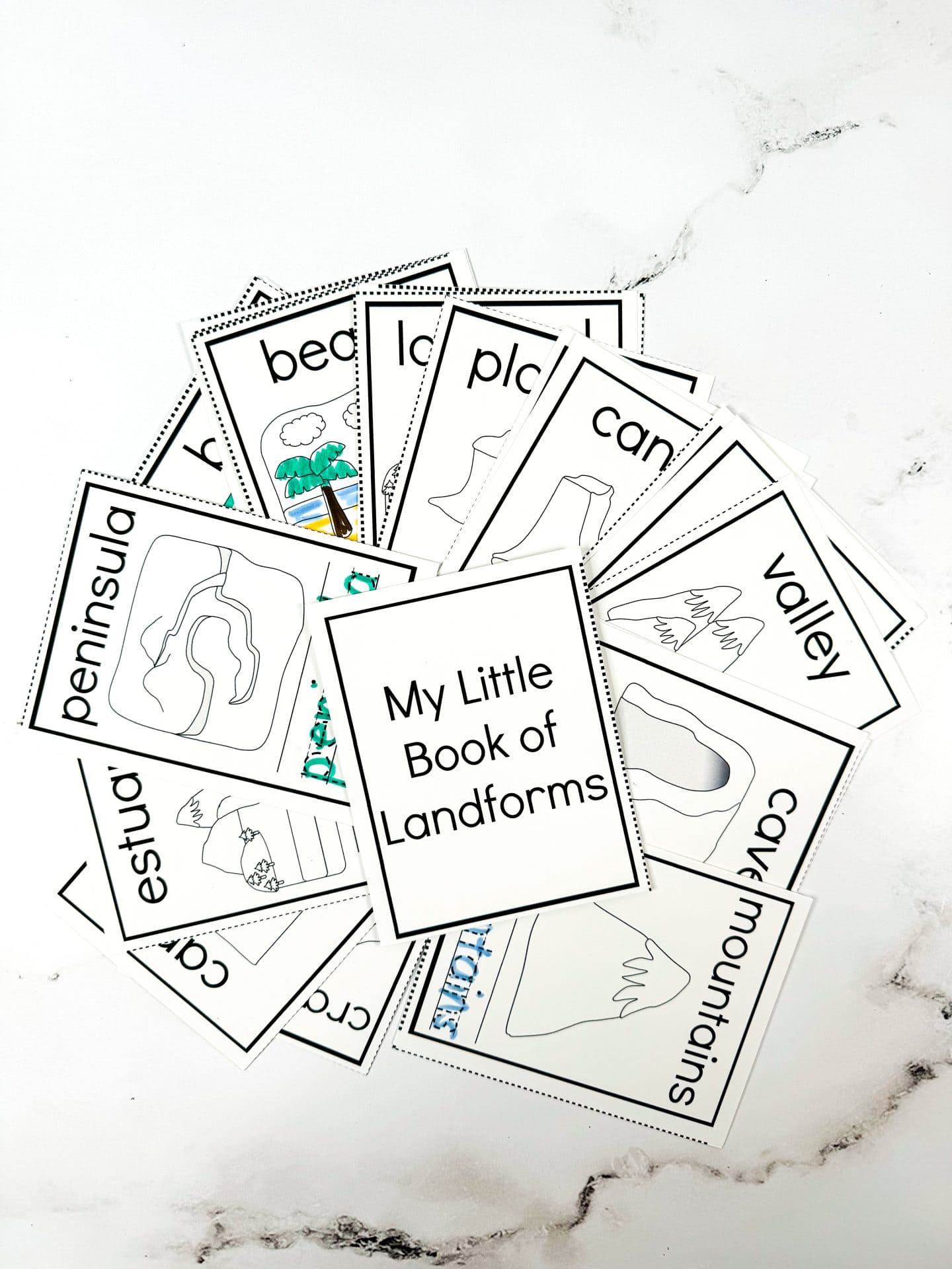 This image shows a fanned-out set of black-and-white landform flashcards titled “My Little Book of Landforms.” The cards feature simple line illustrations and bold labels for various landforms including beach, plateau, canyon, valley, cave, mountains, peninsula, estuary, and crater lake. A few are partially colored in, making it clear this set is designed for hands-on learning and coloring fun.
