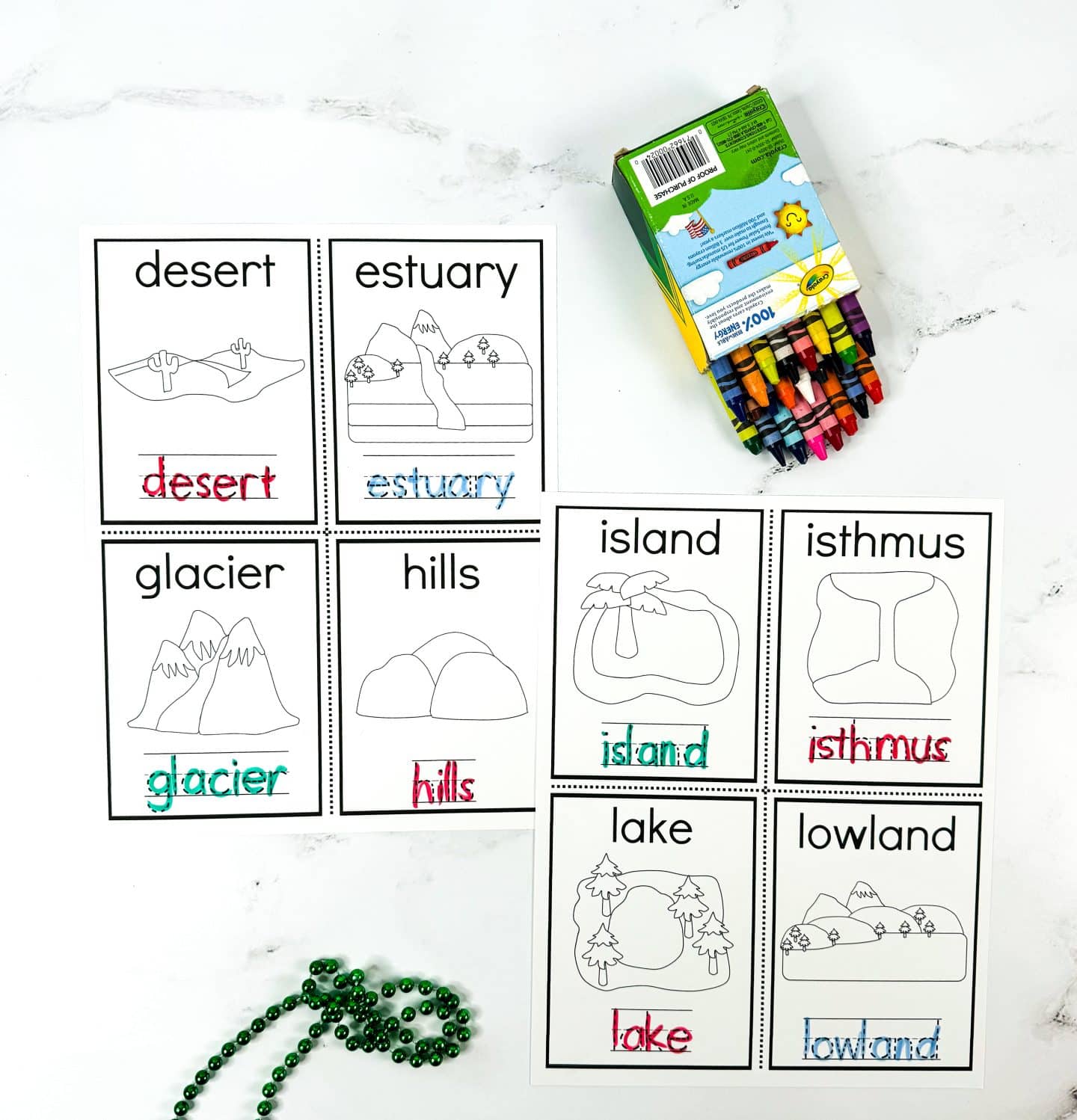 This image shows a colorful set of printable landform flashcards titled “My Little Book of Landforms.” Each card features a simple black-and-white illustration, the landform name, and a version of the name traced in colored marker. Visible landforms include: desert, estuary, glacier, hills, island, isthmus, lake, and lowland. A box of Crayola crayons and a green bead necklace sit nearby, reinforcing the playful, hands-on learning vibe.

