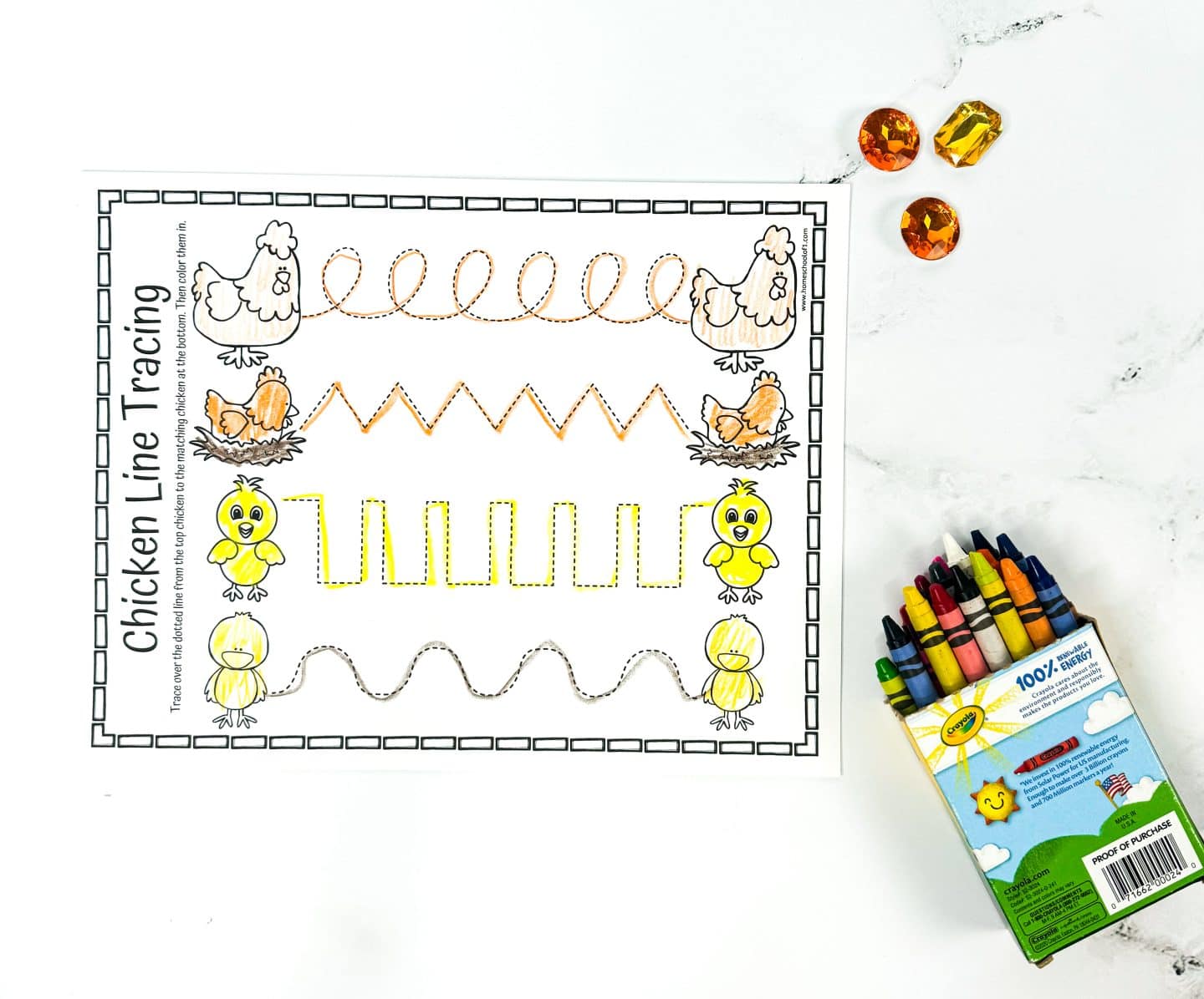 A chicken-themed line tracing worksheet with various zigzag and curved patterns in orange, green, and black. The worksheet is accompanied by a box of crayons and three amber-colored gems on a white surface.
