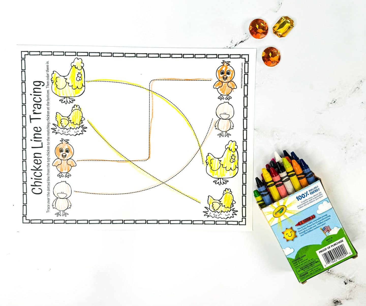 A chicken line tracing worksheet with various curved patterns in orange and yellow. The worksheet is accompanied by a box of crayons and three amber-colored gems on a white surface.
