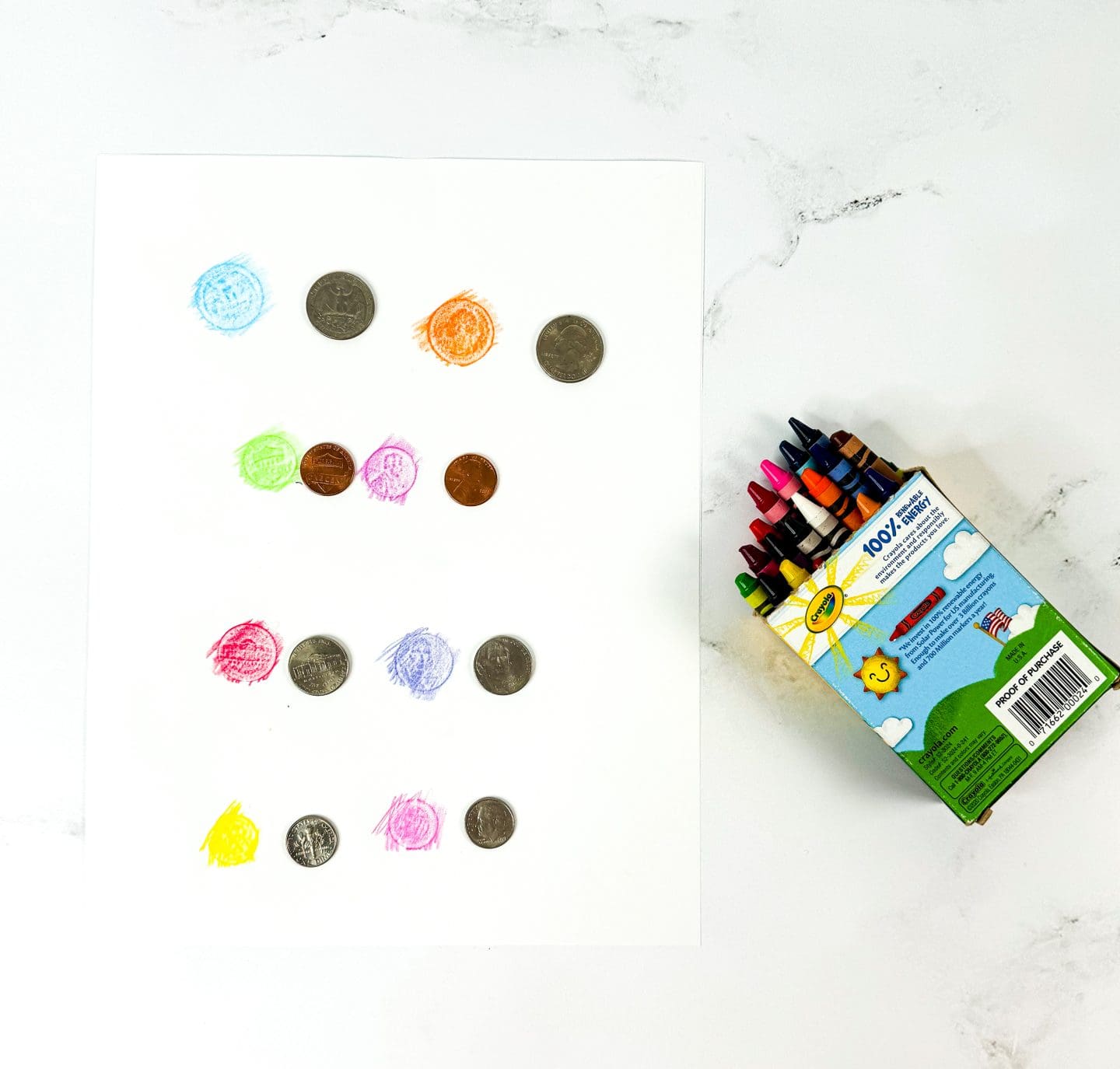 A white sheet of paper displays coin rubbings in crayon, with each coin matched to a colorful rubbing beside it. The coins include pennies, nickels, dimes, and quarters, and each rubbing uses a different crayon color like blue, orange, green, red, and pink. To the right of the paper is an open Crayola crayon box with crayons sticking out, resting on a white marble surface.