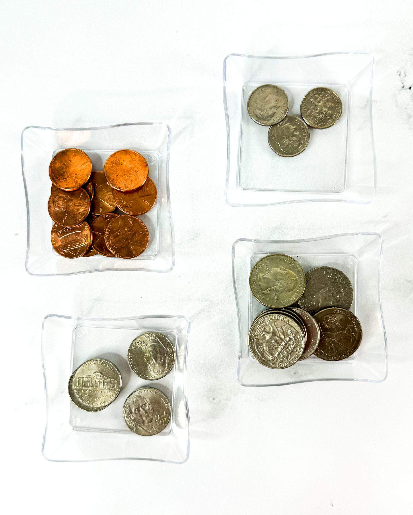 Four clear square containers hold different types of U.S. coins on a white surface. The top left holds a pile of shiny copper pennies, the top right has a few nickels, the bottom left has three Jefferson nickels, and the bottom right contains several quarters, including some with state or commemorative designs