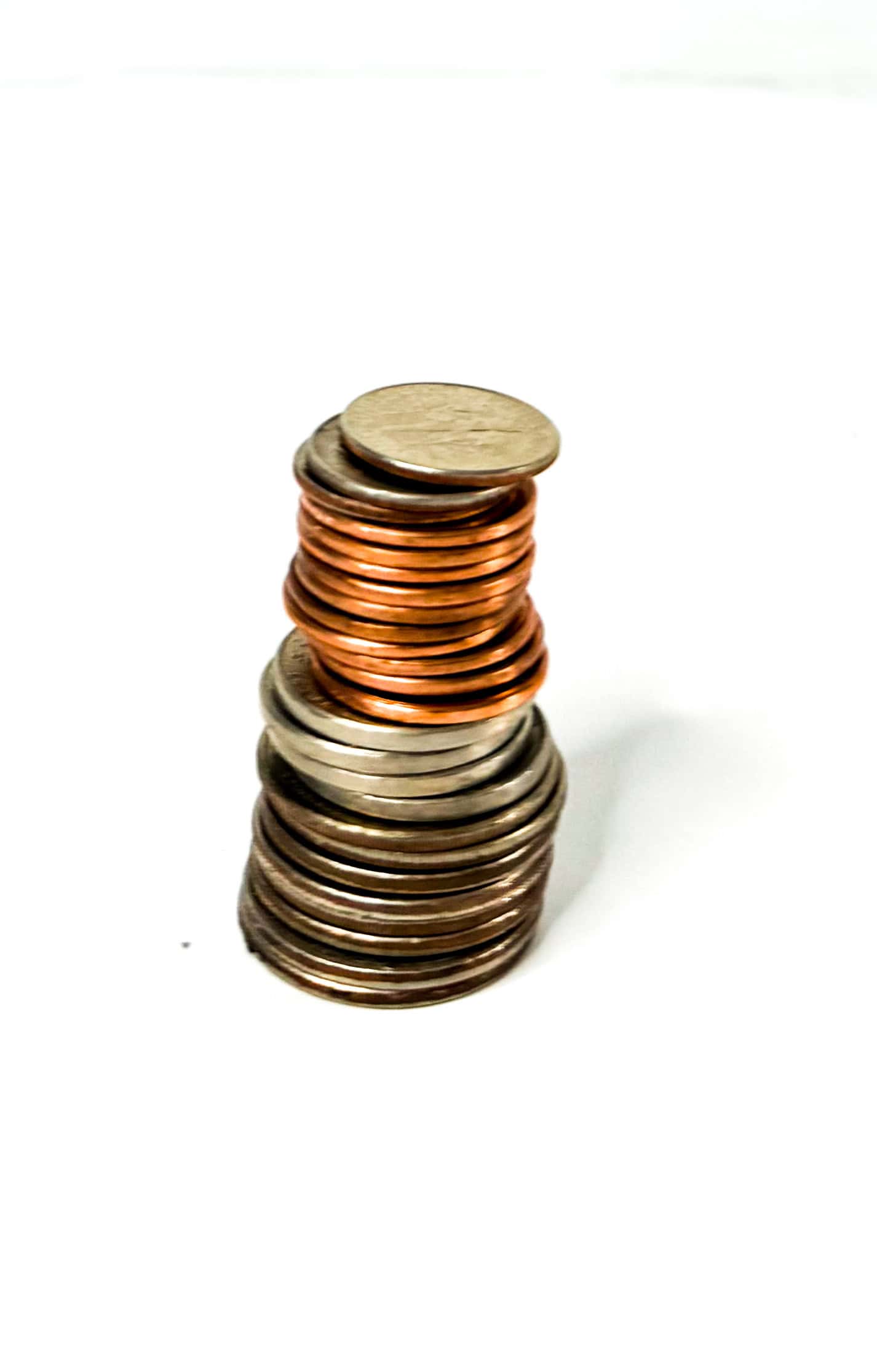 A slightly leaning stack of assorted U.S. coins, including copper pennies and silver-toned nickels and dimes, set against a plain white background. The coins are loosely aligned, giving the stack a wobbly, unsteady appearance.