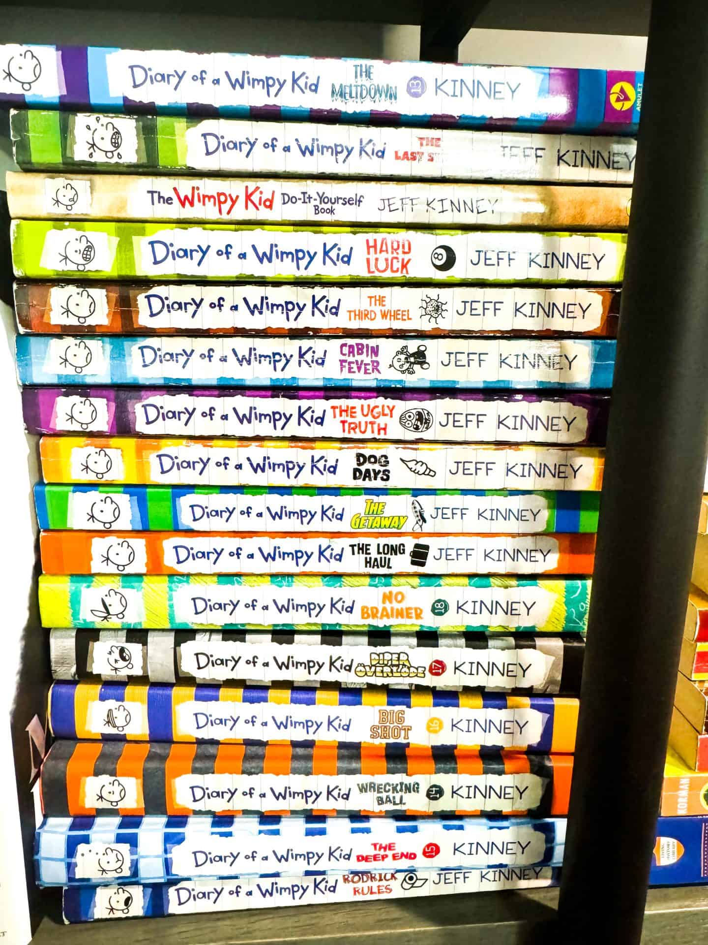 A colorful stack of Diary of a Wimpy Kid books by Jeff Kinney is arranged horizontally on a bookshelf. Titles include The Meltdown, The Last Straw, Cabin Fever, The Long Haul, Big Shot, Wrecking Ball, and Rodrick Rules, among others, showcasing the variety and popularity of the series.

