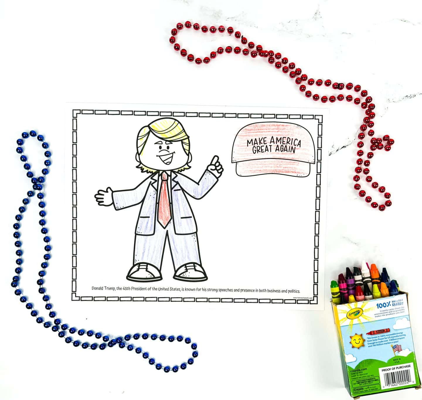A completed coloring worksheet shows a cartoon version of Donald Trump in a suit with a red tie and a speech bubble featuring a red hat that says Make America Great Again. The caption at the bottom describes him as the 45th President of the United States, known for his strong speeches and business background. Red and blue bead necklaces and a box of Crayola crayons are placed around the page for a patriotic classroom vibe.