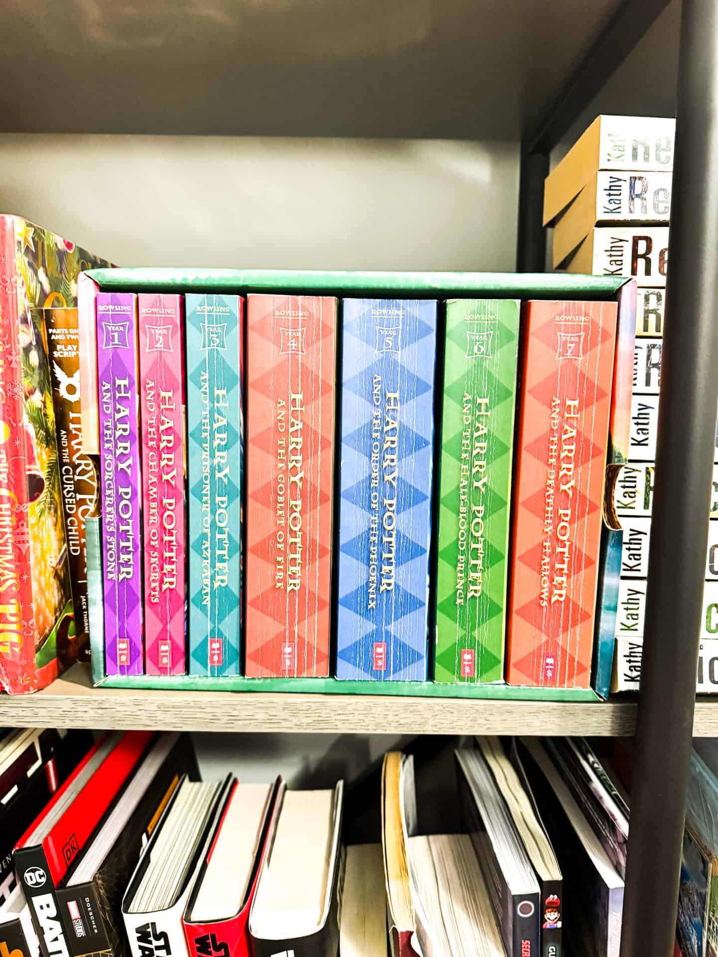 A boxed set of the complete Harry Potter book series by J.K. Rowling is displayed on a bookshelf, with each hardcover volume in a brightly colored cover—ranging from purple and red to blue and green. The set is neatly arranged in order from The Sorcerer’s Stone to The Deathly Hallows, surrounded by other books including Harry Potter and the Cursed Child and various titles by Kathy Reichs.

