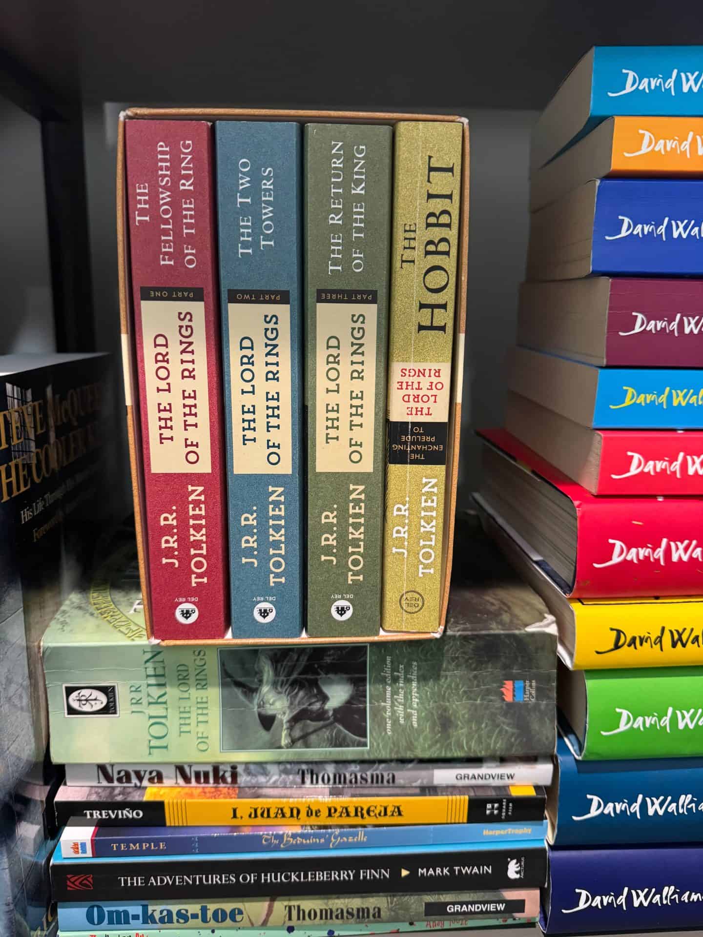 A boxed set of J.R.R. Tolkien's The Lord of the Rings trilogy and The Hobbit sits on a bookshelf. The spines display The Fellowship of the Ring, The Two Towers, The Return of the King, and The Hobbit, each in earth-toned covers. Surrounding the set are additional books including David Walliams novels, The Adventures of Huckleberry Finn by Mark Twain, and several titles by Thomasma and other classic and historical fiction authors.

