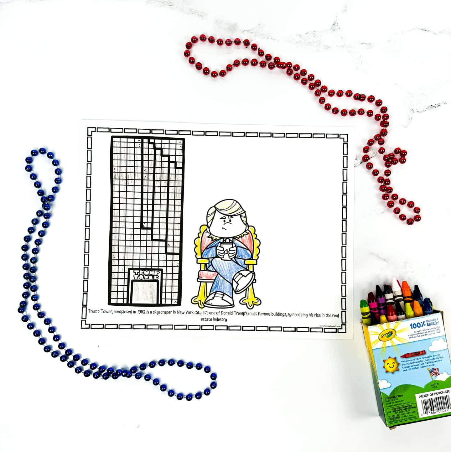 A completed coloring worksheet features a cartoon Donald Trump sitting in a gold chair next to a drawing of Trump Tower, with the building partially colored in black and labeled at the base. A caption at the bottom explains Trump Tower’s completion in 1983 and its significance in Trump’s real estate career. Red and blue beaded necklaces and a box of Crayola crayons are arranged around the page.